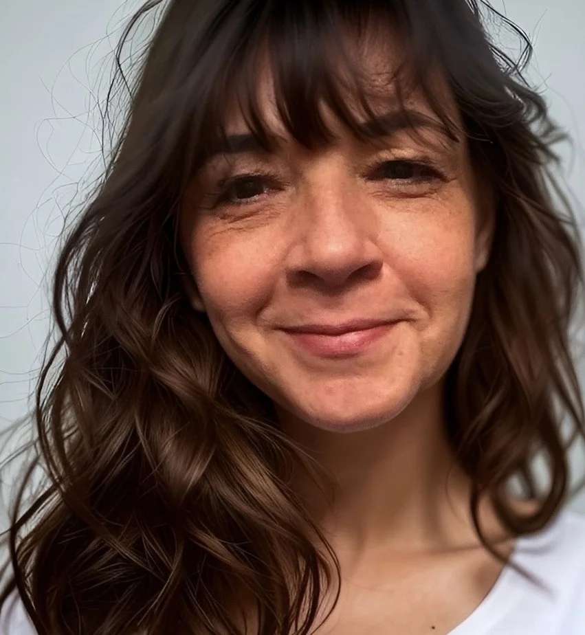 Close-up of a woman with brown hair, smiling softly, wearing a white top, in a well-lit environment.