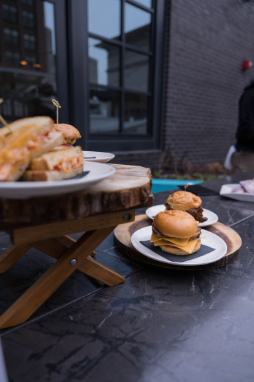 Two plates of sliders on a black table, one with small cheeseburgers and the other with mini sandwiches, set outdoors near a modern building with large windows.