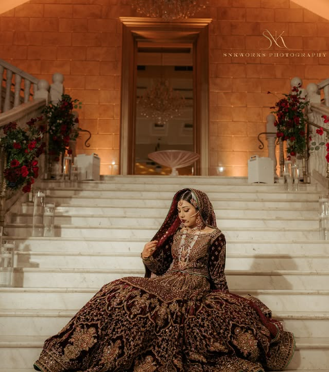 A woman in traditional South Asian wedding attire sitting on the stairs of a grand interior venue decorated with floral arrangements.