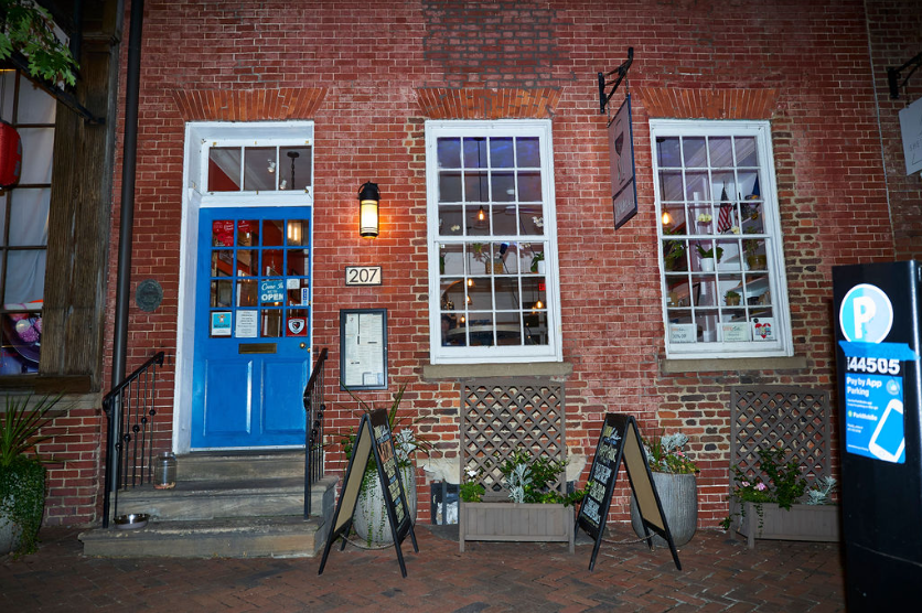 A brick storefront with a bright blue door, three large windows, chalkboard signs, and potted plants outside. The address “207” is displayed next to the door—photographed by OMGVA, a Virginia-based video production company serving DC and the DMV.