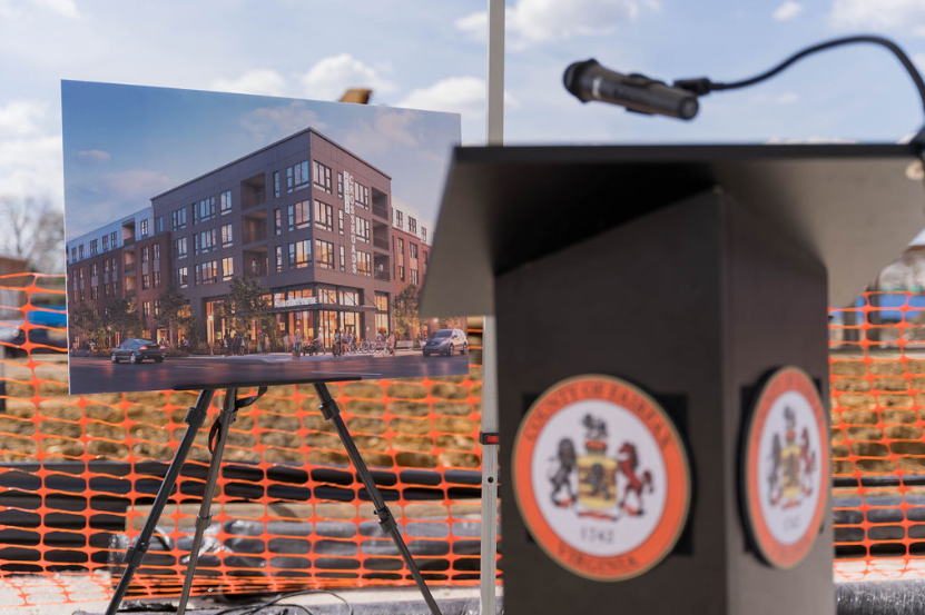 A construction site features a rendering of a modern apartment building on an easel and a podium with the Maryland state seal, with orange safety fencing in the background.