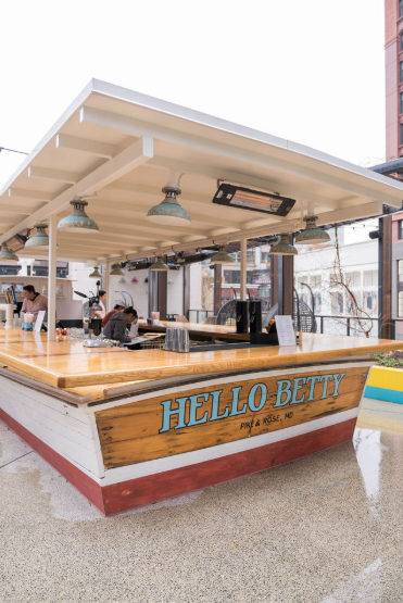 Outdoor eatery bar with a wooden counter beneath a white awning, featuring a sign that reads "HELLO BETTY, MUKE & ROLLS, MO," with ceiling lights and fans—captured by OMGVA, a full-service video production company based in Virginia and serving Washin