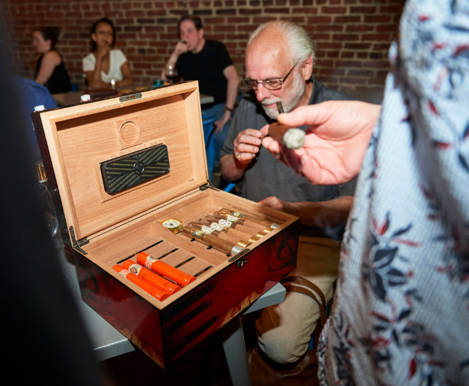 A group of people gathered around a table at a social event, featuring a red wooden cigar box with cigars and a cutter, as one person holds a lit cigar—captured by OMGVA, a Virginia-based video production company serving Washington DC and the DMV.