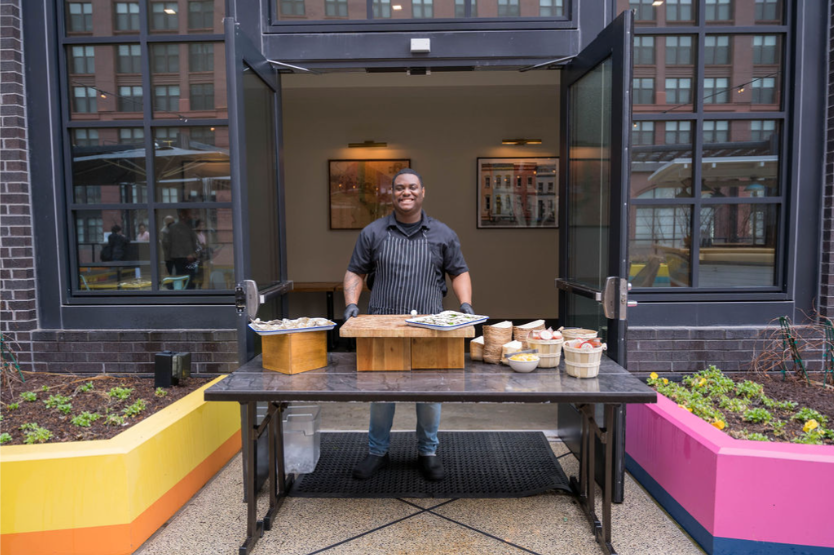 A smiling man in a black shirt and striped apron stands behind a stand with bowls and plates, outdoors near a modern building with large windows, with colorful planters beside him.