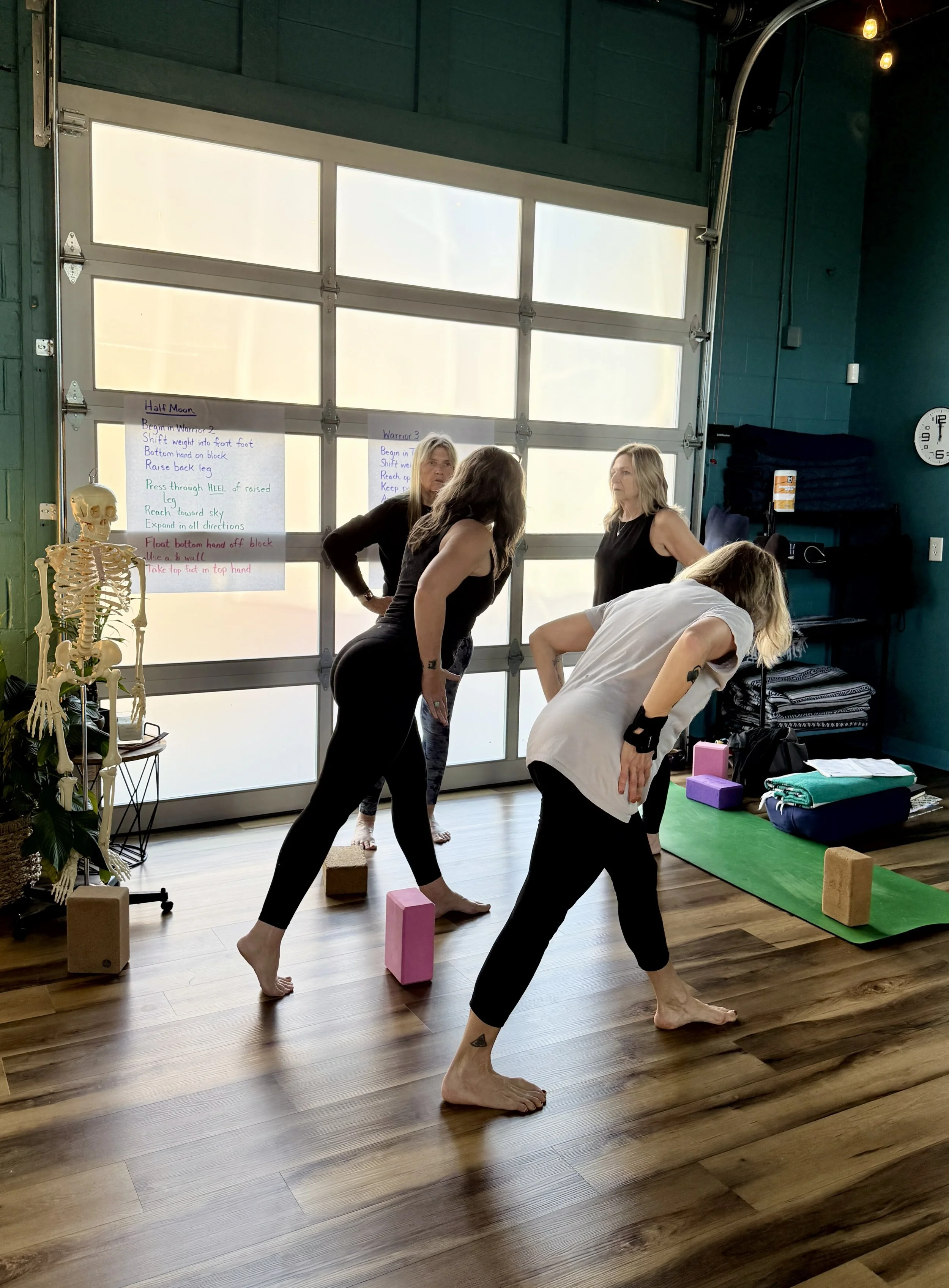 Four women participating in a yoga or stretching class indoors, using yoga blocks and mats, with a skeleton model and instructional posters in the background.
