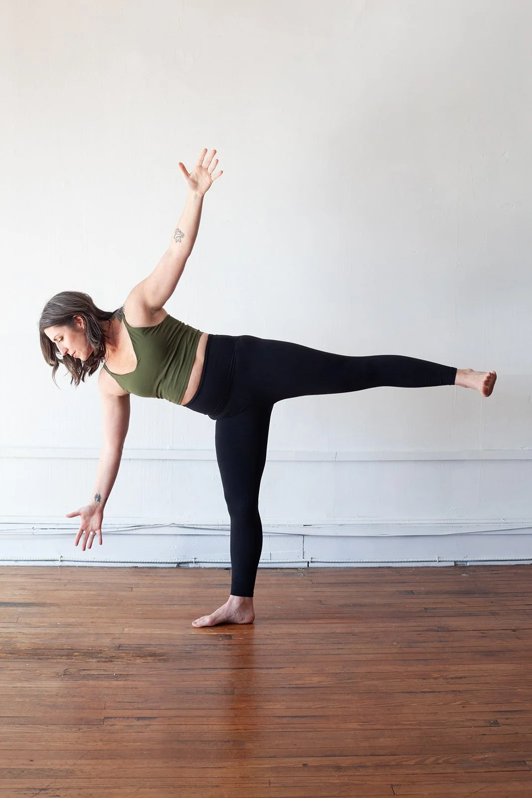 Woman practicing yoga indoors on a wooden floor, balancing on one leg with her right arm extended downward and her left arm raised upward, wearing a green sports bra and black leggings.