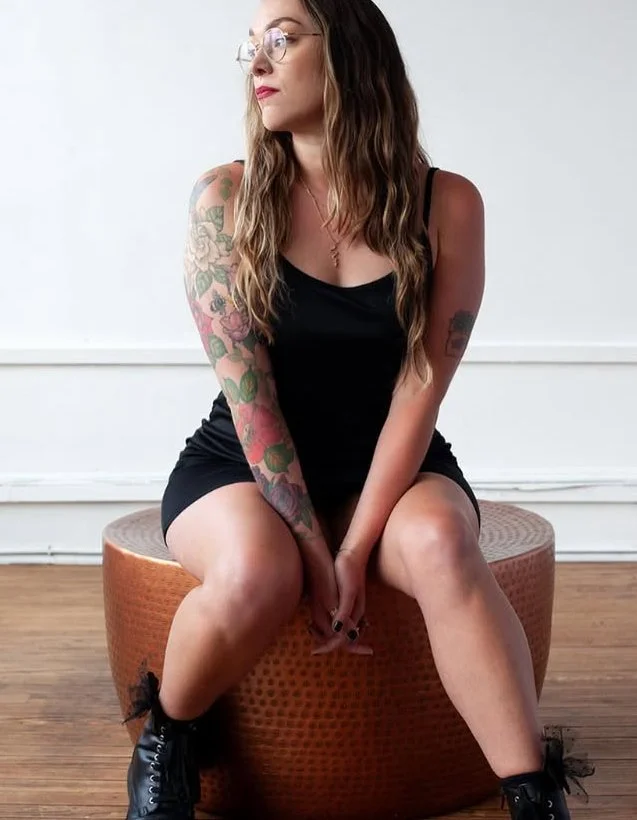 A young woman with long wavy hair, wearing glasses, a black sleeveless dress, and combat boots, sitting on a round padded stool with her hands clasped between her knees.