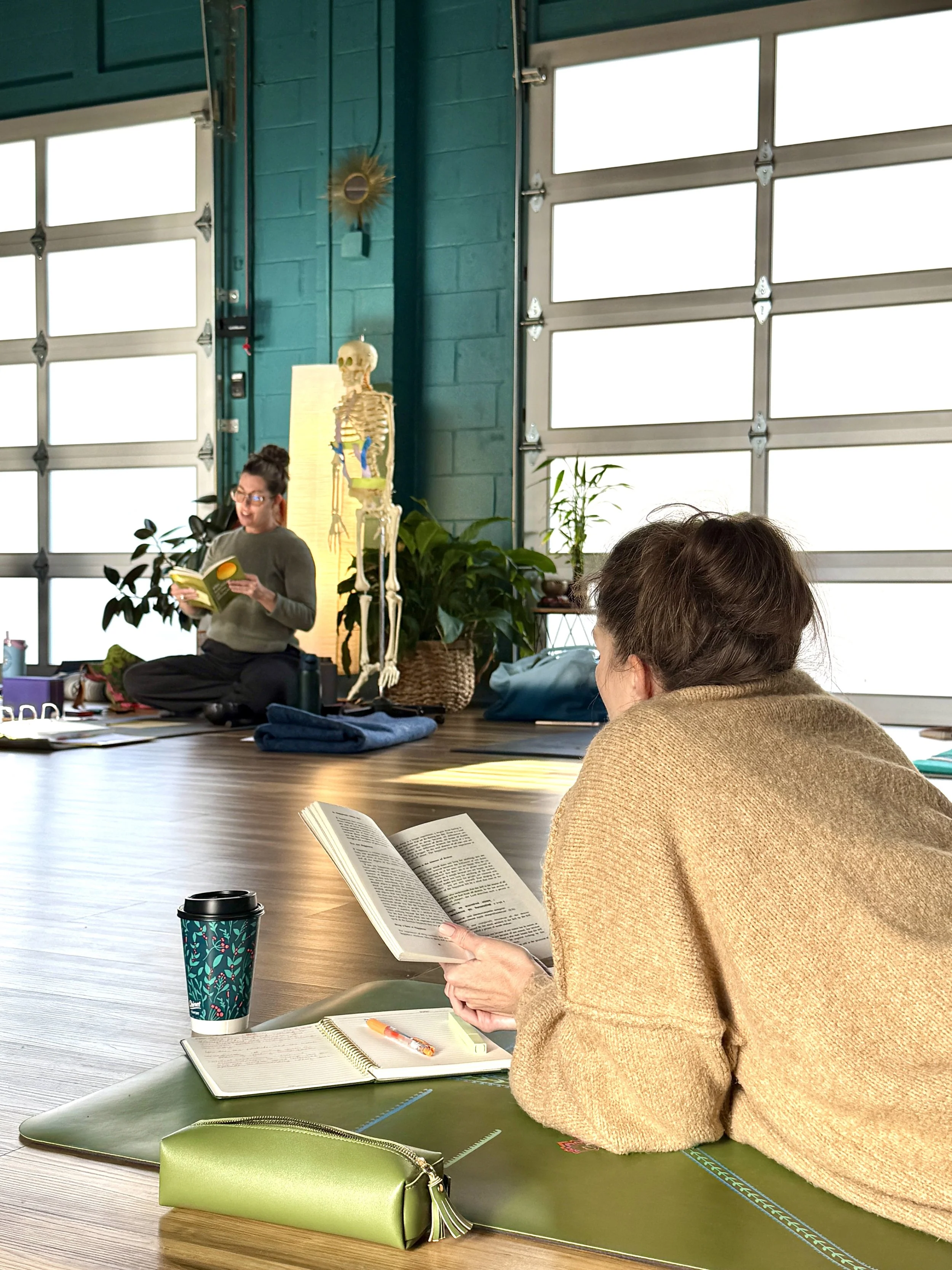A woman in a brown sweater reading a book, sitting on a yoga mat at a table in a bright room with large windows, a skeleton model, and green plants.