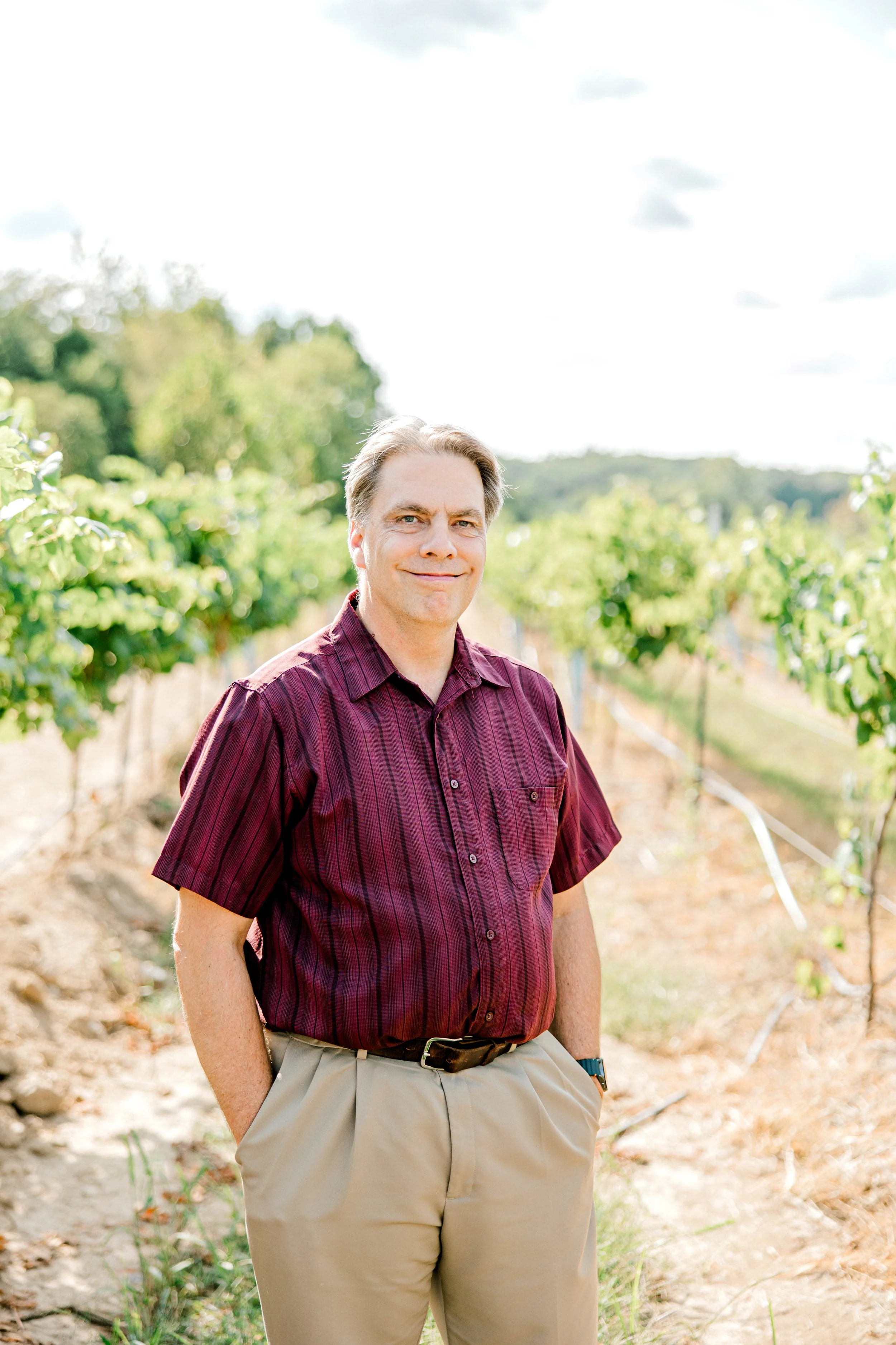 brown haired man wearing a striped maroon shirt smiling and standing in a vineyard