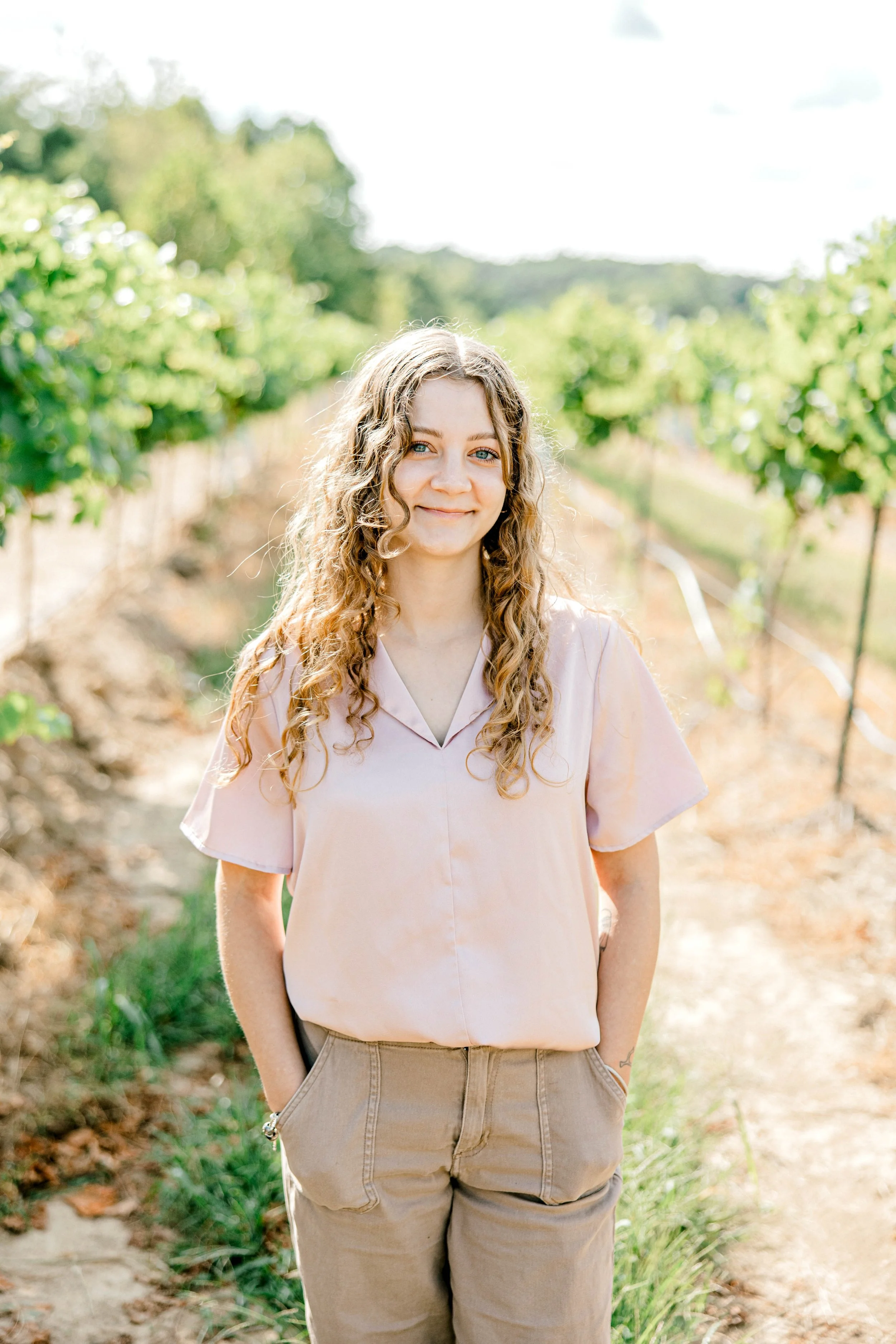 young woman with long curly hair smiling and standing in a vineyard