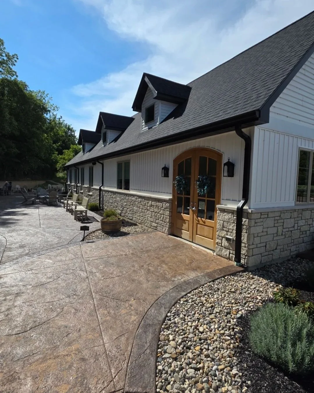 image of a white and stone building with a black roof and large wooden doors, adjacent to a patio and stamped concrete walkway with landscaping and blue skies overhead