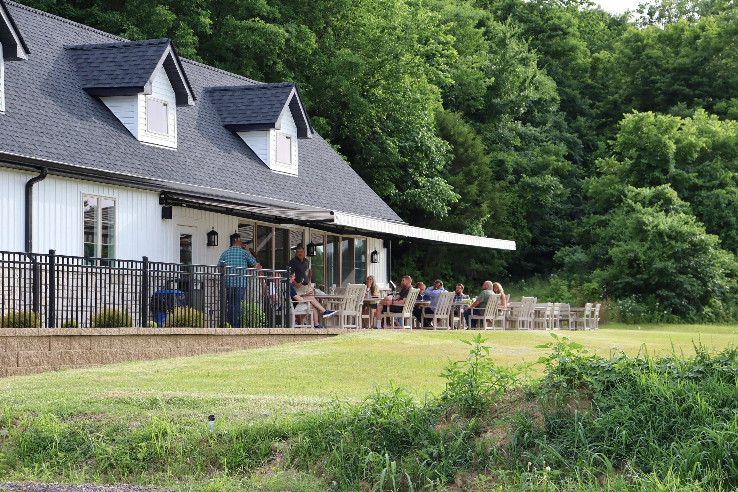 people seated on outside patio at a white building with a black roof near woods