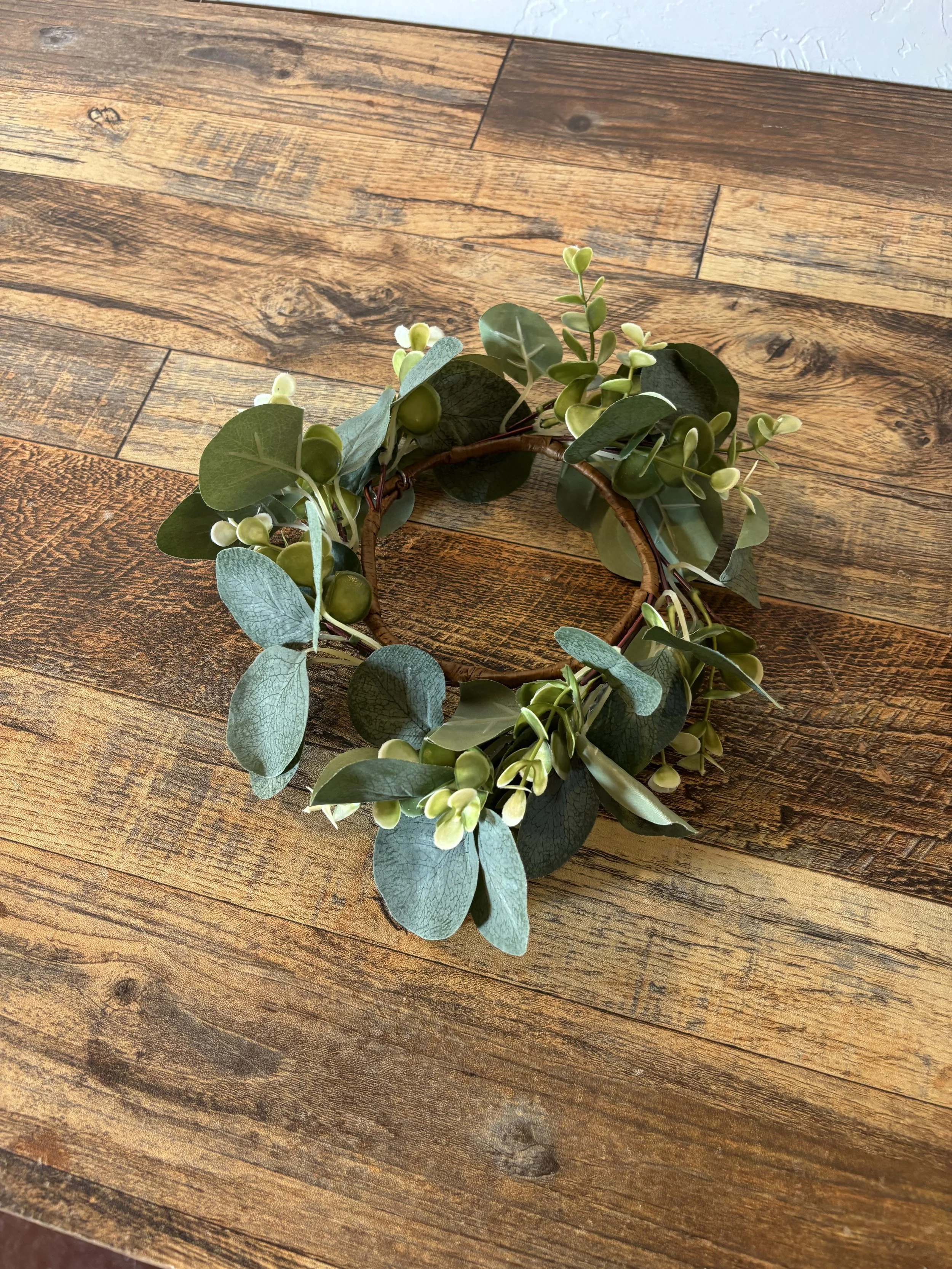Round floral wreath made of green leaves and small white buds on a wooden table.