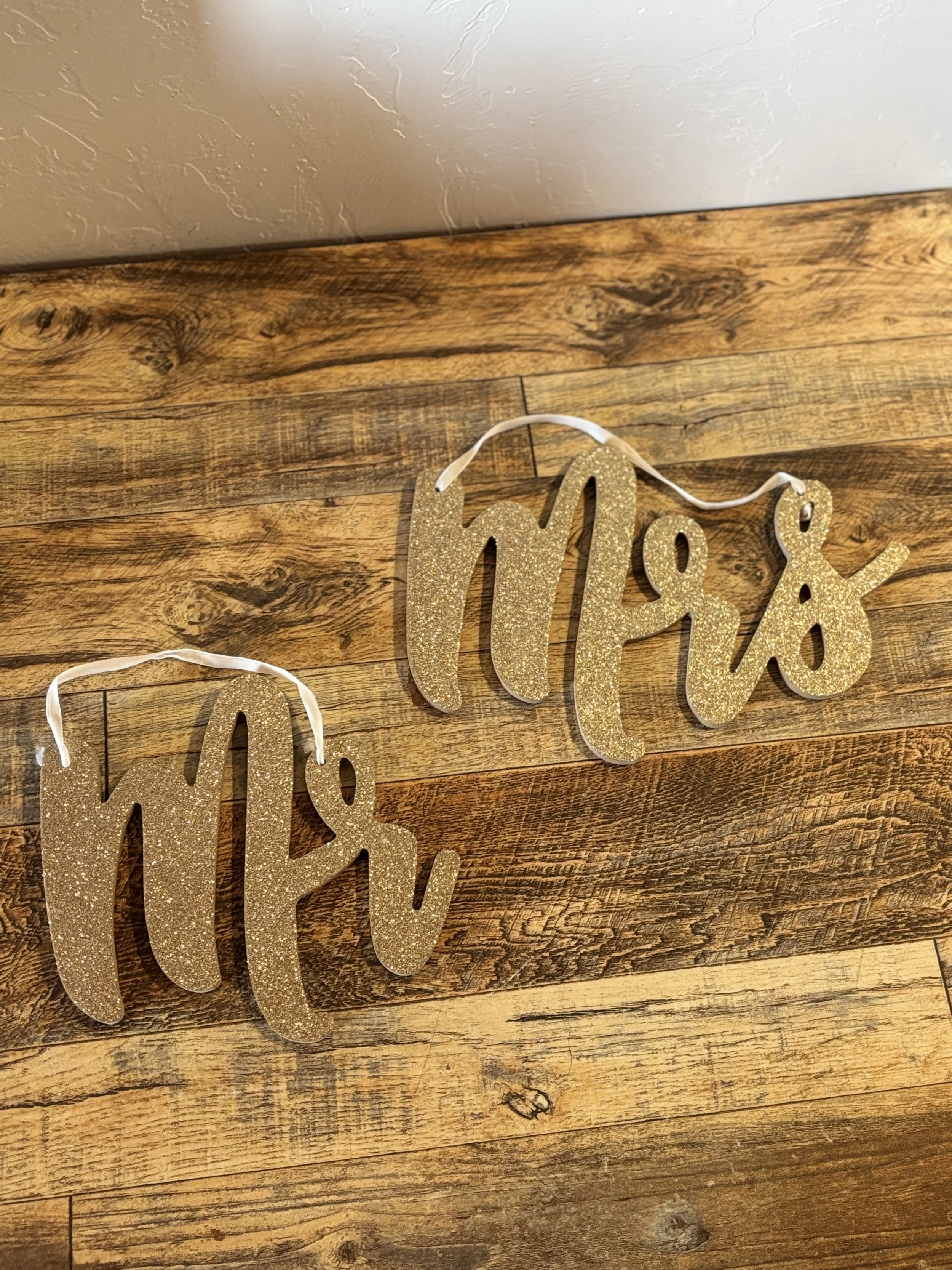 Decorative gold glitter 'Merry' signs hanging with white ribbons on a wooden surface.