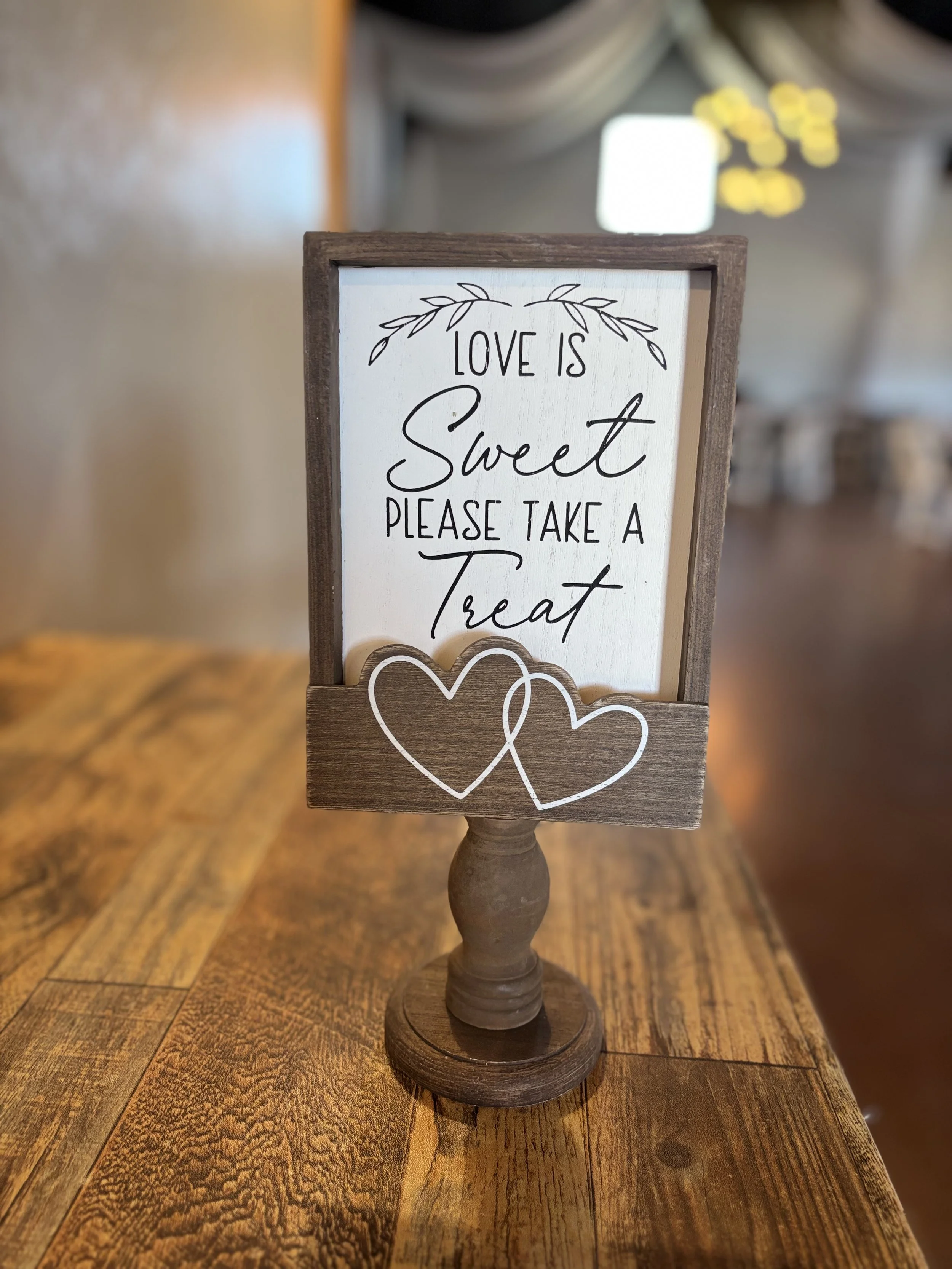 A decorative tabletop sign with a wooden base and frame. The sign reads "Love is Sweet Please Take a Treat" in black handwritten-style text, with outline drawings of two overlapping hearts at the bottom. The background of the sign is white, and the setting appears to be indoors with warm lighting.