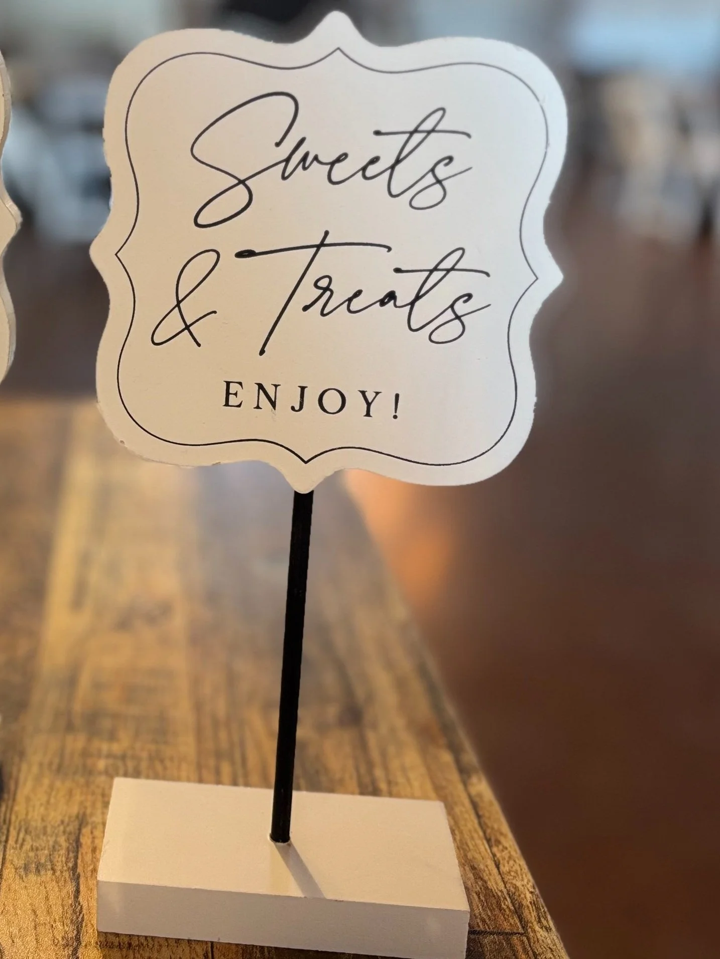 A decorative sign on a stand that reads "Sweets & Treats Enjoy!" placed on a wooden surface.