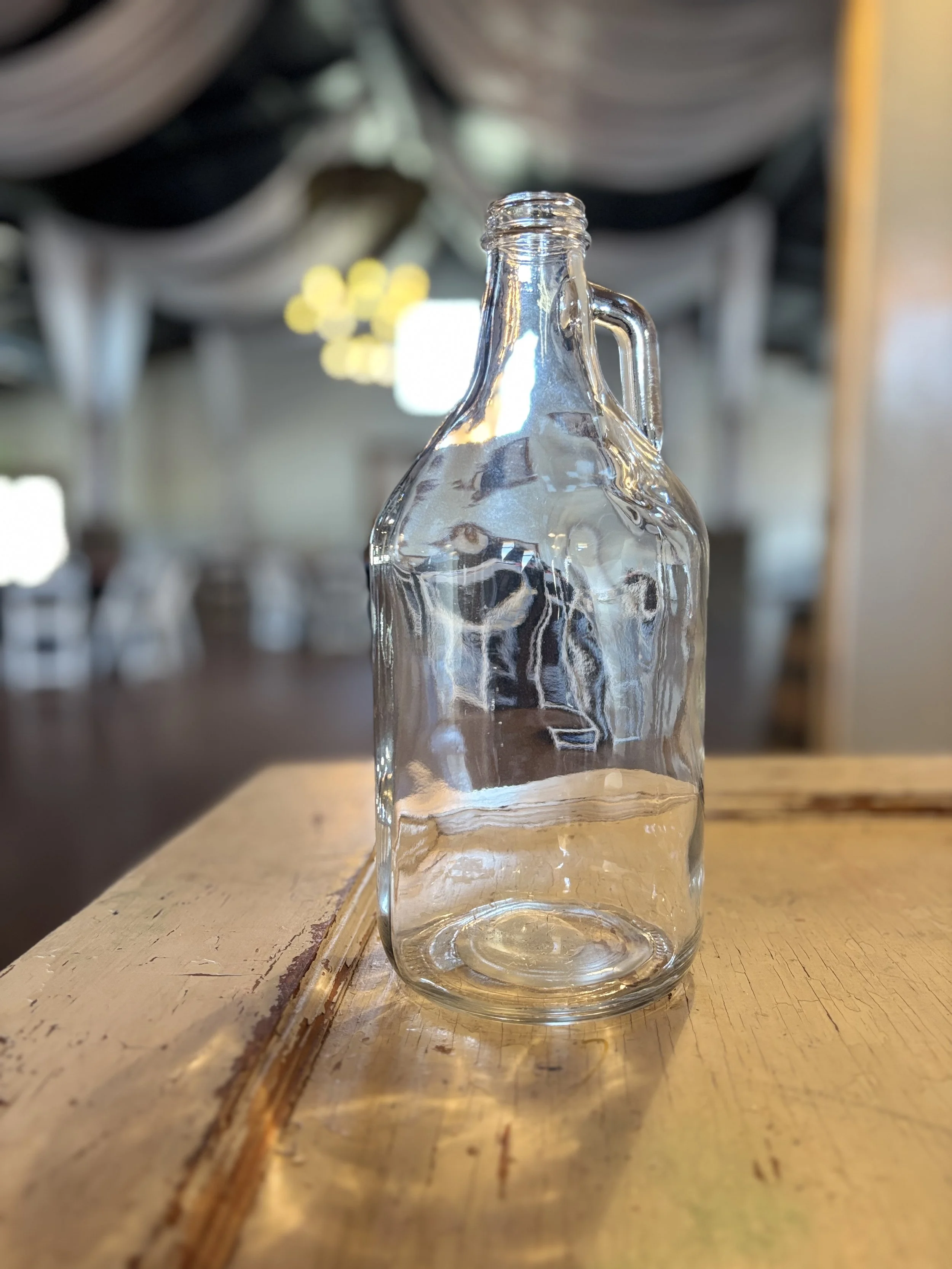 A clear glass bottle with a handle, empty, placed on a wooden table in a restaurant or cafe setting.