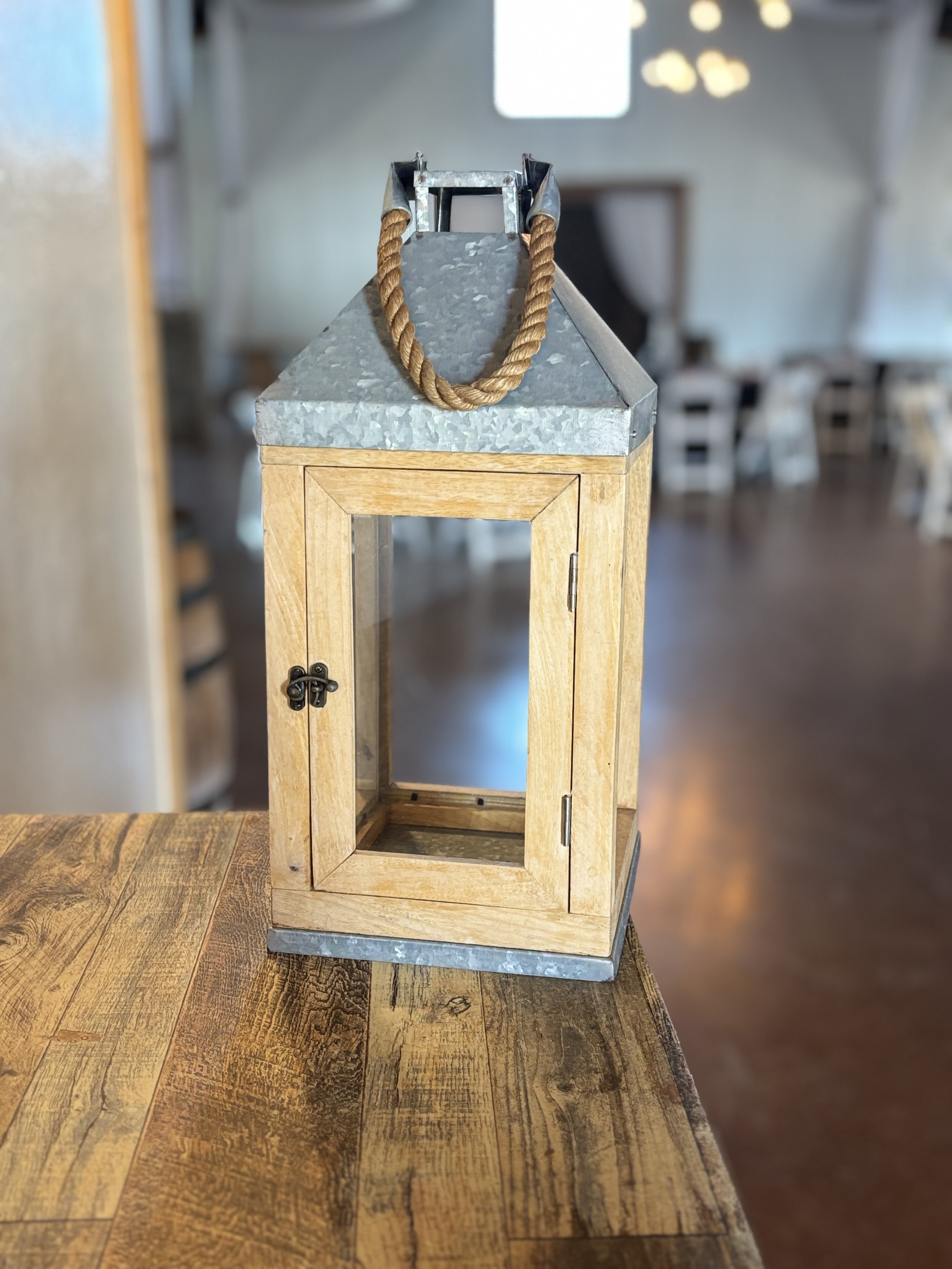 A decorative lantern with a wooden frame, a glass panel, a metal top, and a rope handle on a wooden table inside a rustic room.