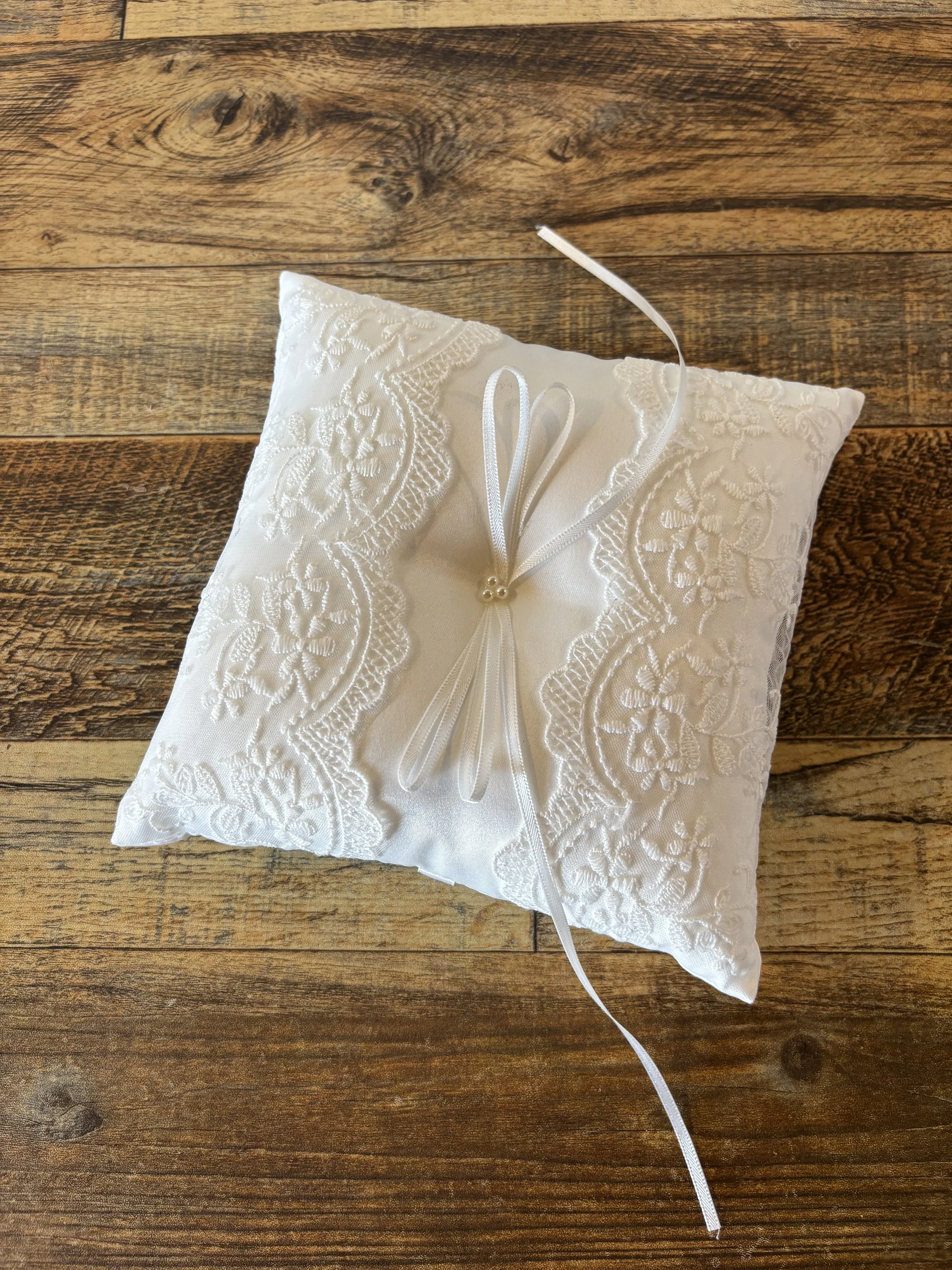 An off-white satin ring pillow with embroidered lace details, decorated with a satin ribbon bow with pearl accents, on a wooden surface.