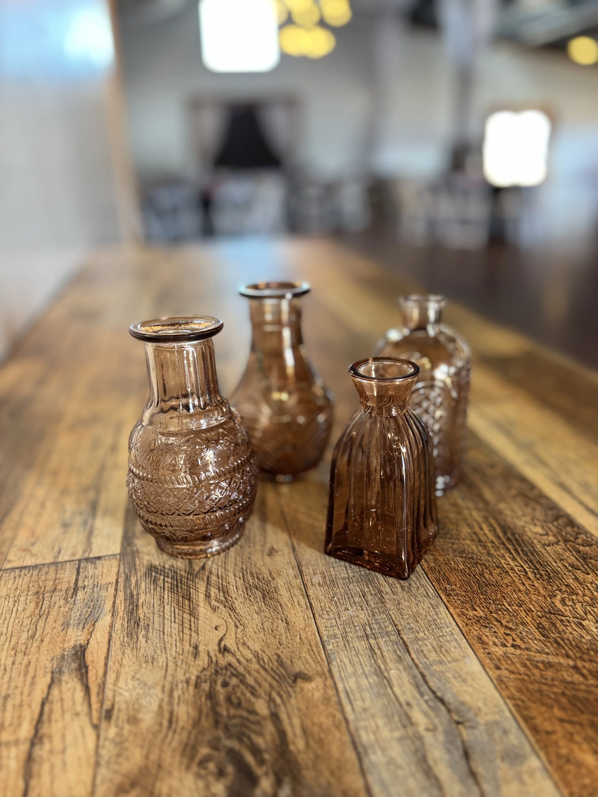 Four small amber-colored glass vases with textured and ribbed patterns placed on a rustic wooden table in a blurred restaurant setting.