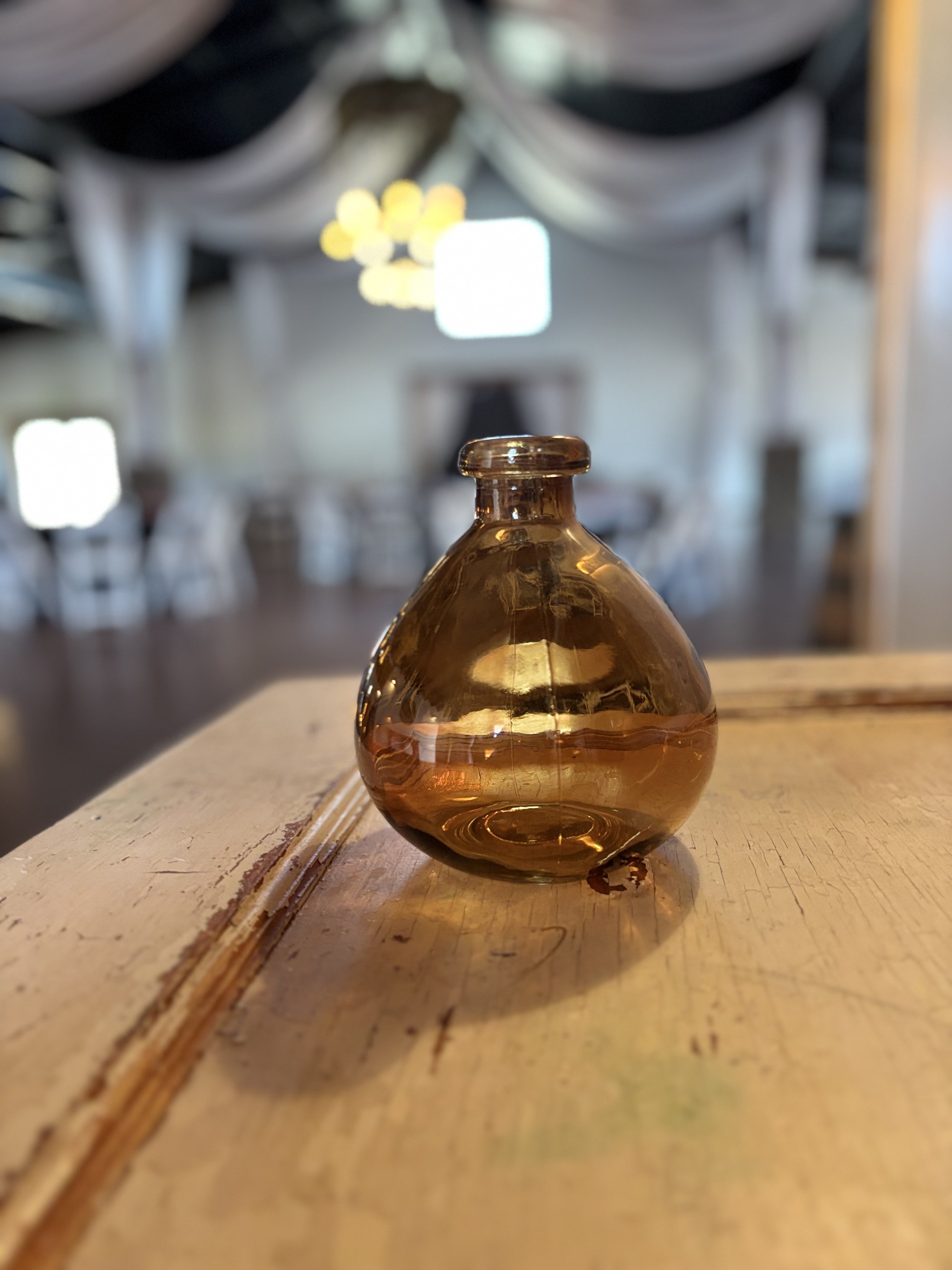 A small amber glass vase on a wooden table in a softly lit room with draped fabric ceiling and blurred chairs and windows in background.