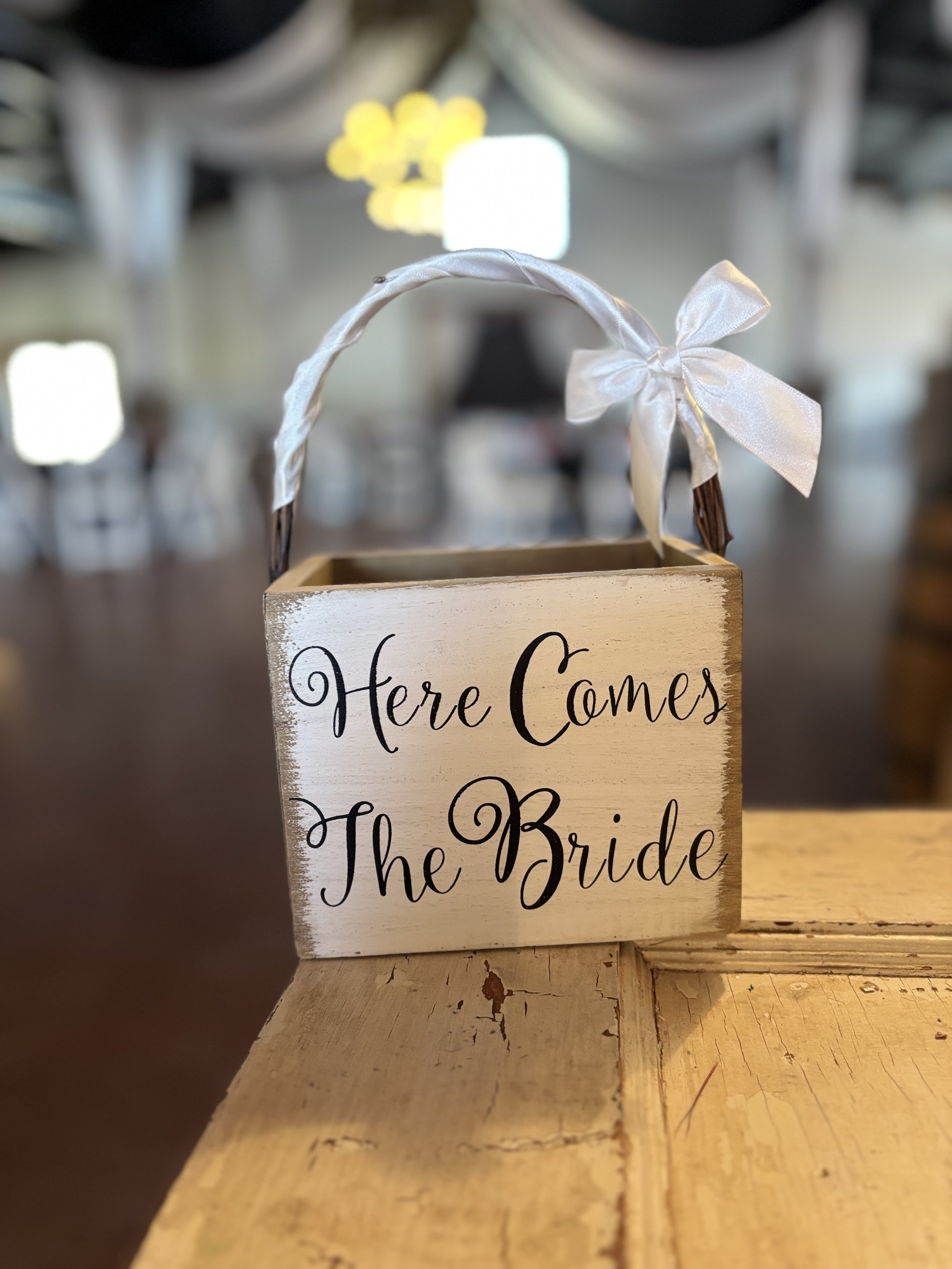 Decorative sign on a wooden table reading "Here Comes The Bride" with ribbon handle and bow, blurred background of an indoor venue.