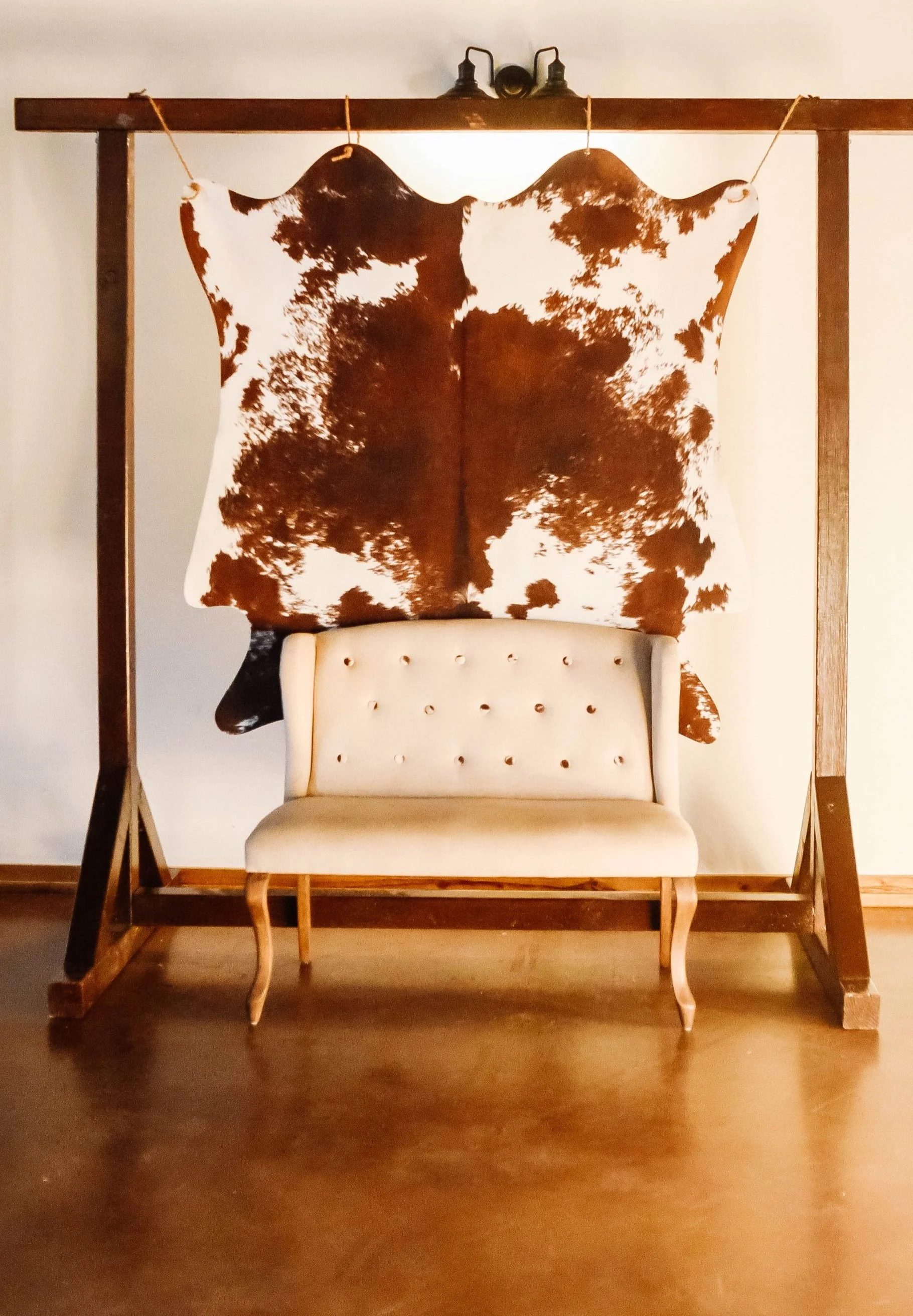 A beige upholstered tufted bench in front of a cowhide decoration hung on a wooden frame. The cowhide has brown and white patterns and is suspended by strings from the wooden frame.