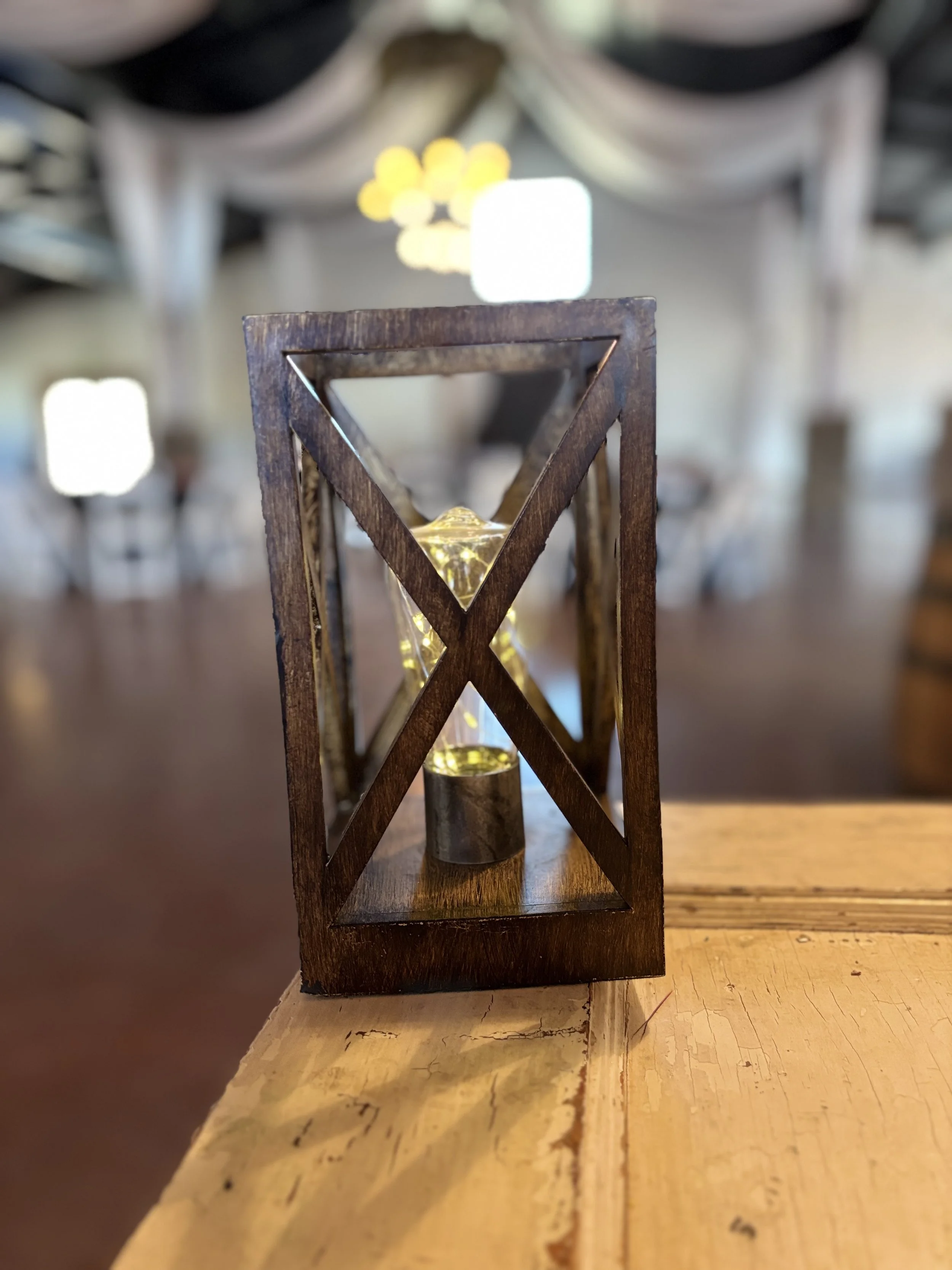 Wooden lantern with a small fairy light inside on a wooden table, blurred background with hanging fabric and lights.