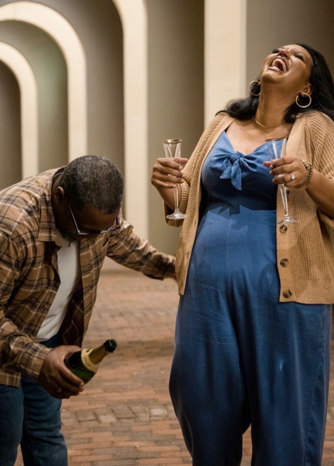 Two people are celebrating in Virginia Beach; a woman with long black hair, earrings, wearing a blue dress and beige cardigan is holding two champagne glasses, laughing joyfully. A man with glasses and short gray hair, dressed in a plaid shirt, is kn