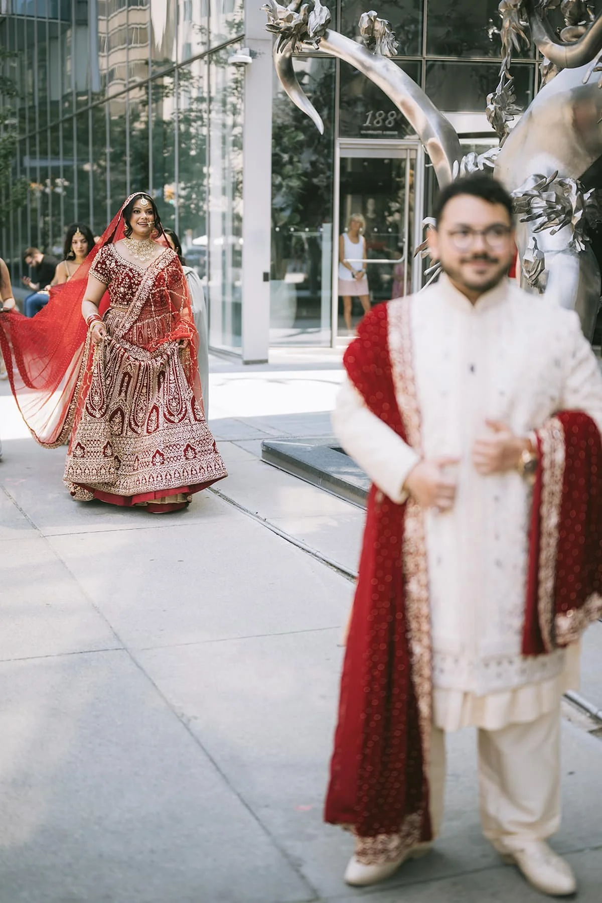 Bride approaches the groom for their first look outside Ricarda's during a modern Hindu wedding in Toronto, wearing a red embroidered lehenga and ivory sherwani.