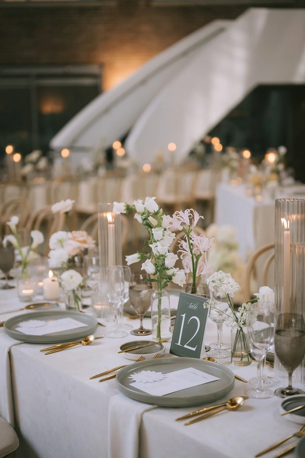 Luxury wedding reception table at Ricarda's Toronto featuring modern bud vase centrepieces, ribbed glass taper candle holders, grey stoneware plates, and gold flatware in a minimalist design.