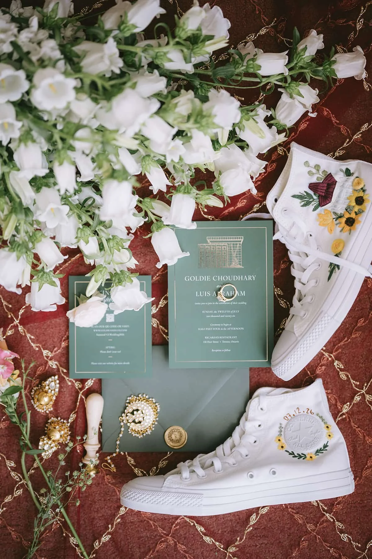 Wedding flat lay featuring custom embroidered Converse sneakers, green wedding invitations, wax seal, jewellery, and white florals for a modern Hindu wedding in Toronto.