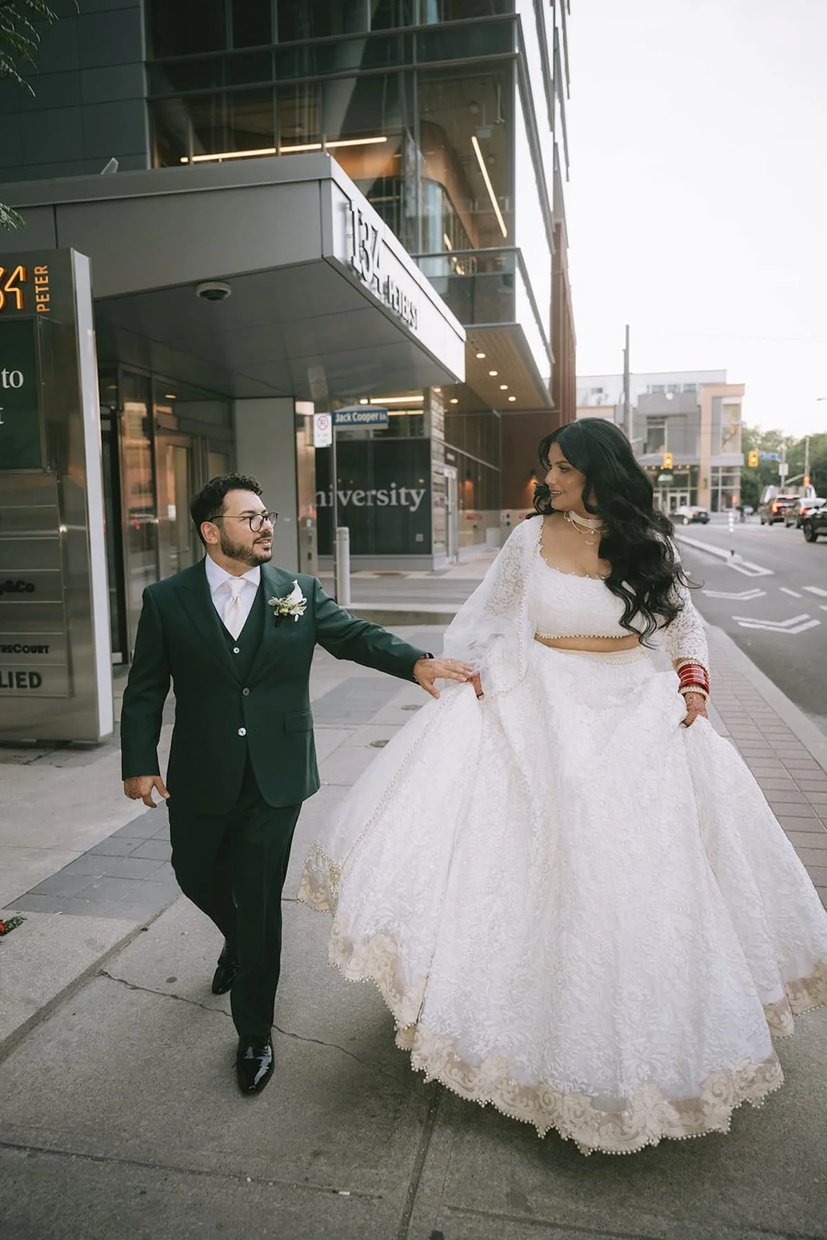 Bride and groom walking hand in hand outside 1 Hotel Toronto after their ceremony, with the bride wearing a modern white lehenga and the groom in an emerald green suit.