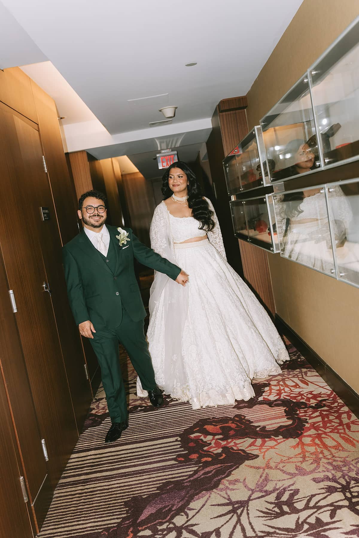 Bride and groom walking hand in hand through the hallway before their ceremony at Ricarda's Toronto, showcasing the bride's modern white lehenga and the groom's emerald green suit.