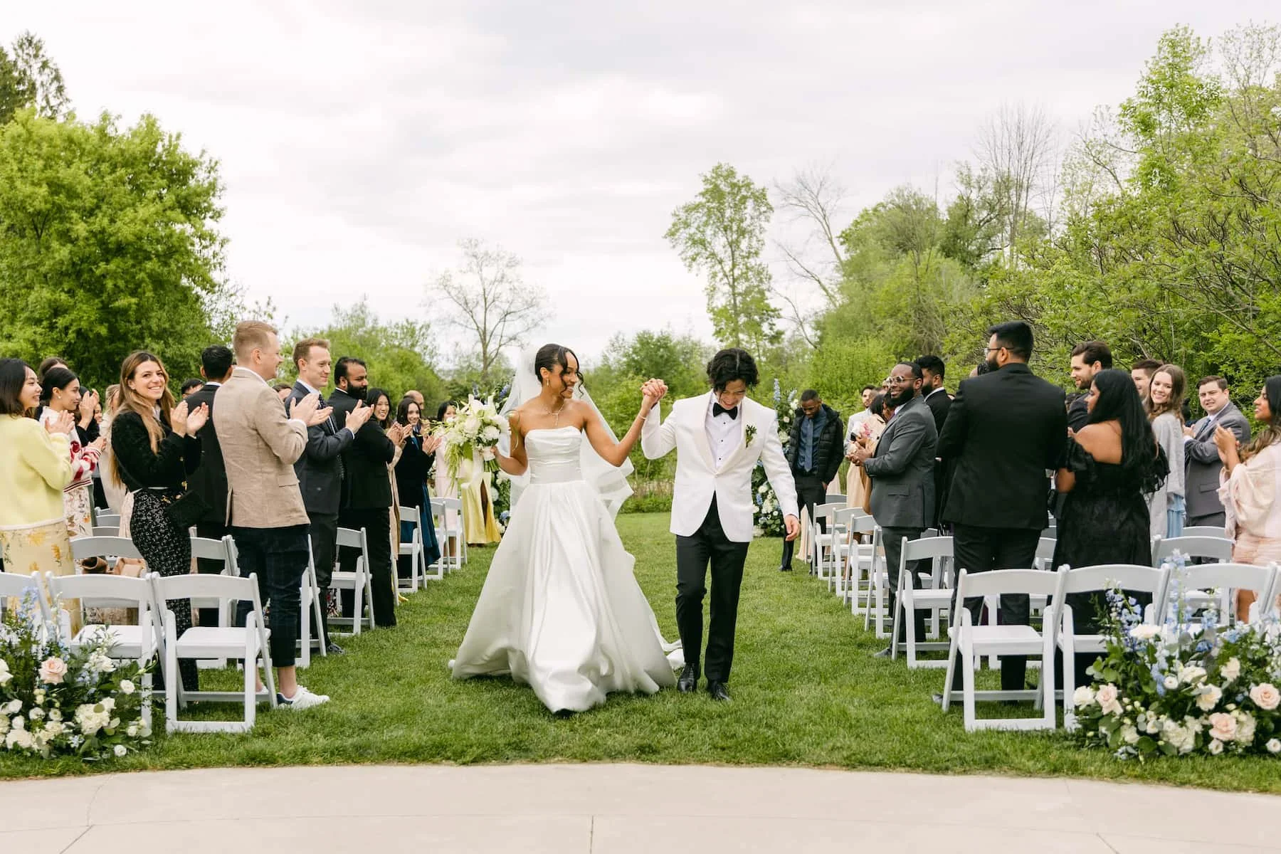 Newly married couple celebrate their ceremony recessional surrounded by cheering guests during an outdoor wedding at Harding Waterfront Estate in Mississauga, Ontario.