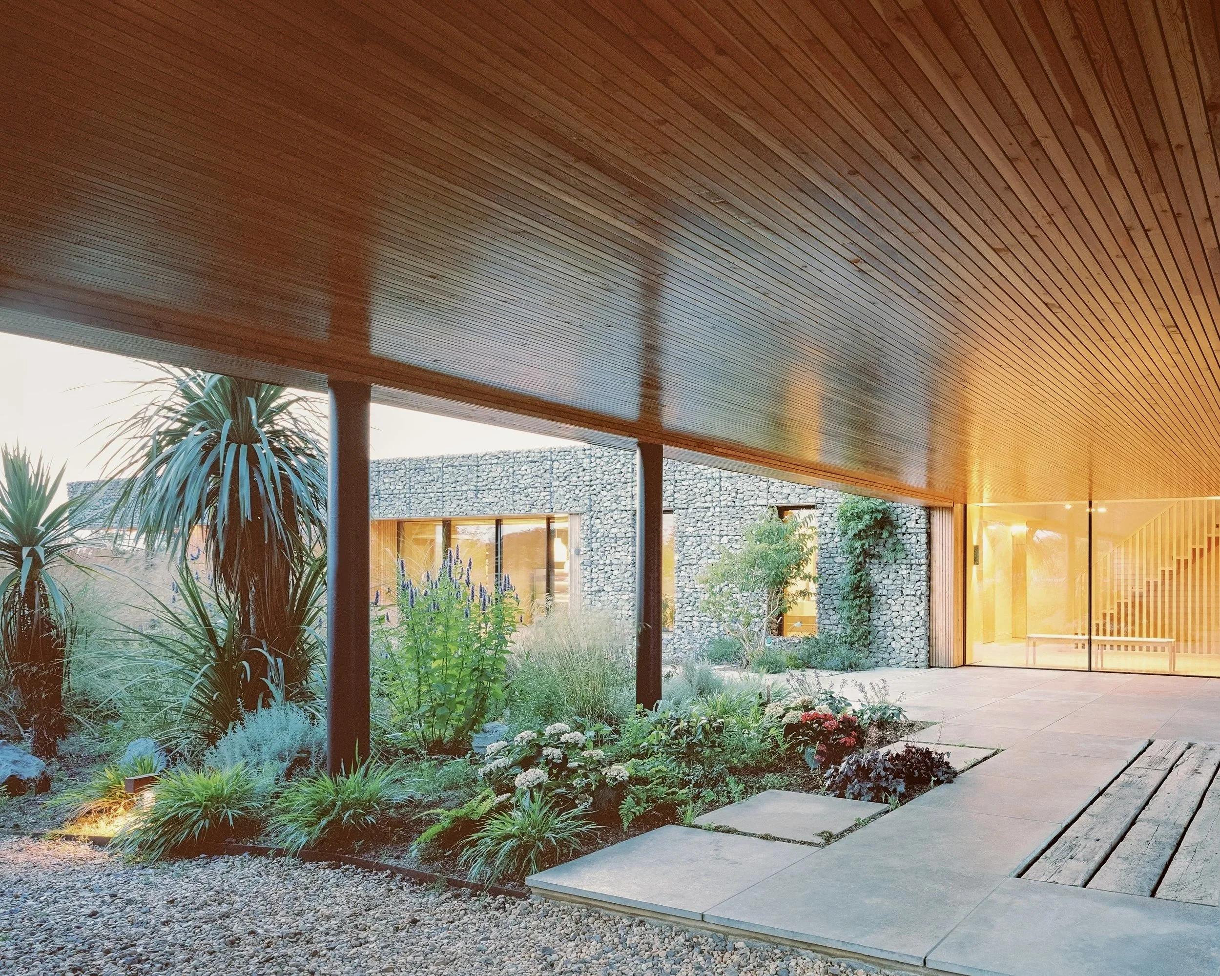 Modern house with large glass windows, stone walls, and a landscaped garden with plants.