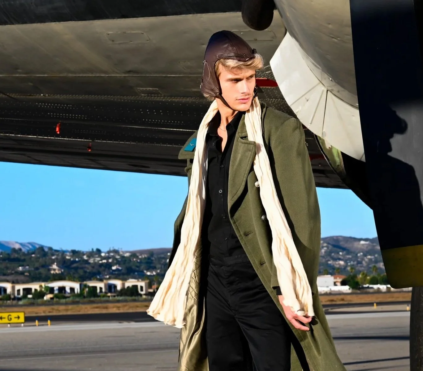 A young man standing next to an aircraft with a woman’s scarf and a leather flying helmet, wearing a long green coat and black clothing.
