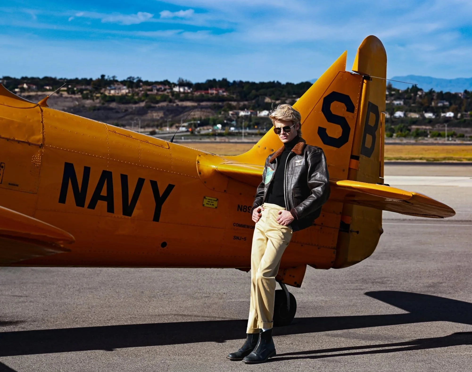 A woman in sunglasses and a leather jacket leaning against a yellow navy aircraft on a runway with a landscape of hills and houses in the background.