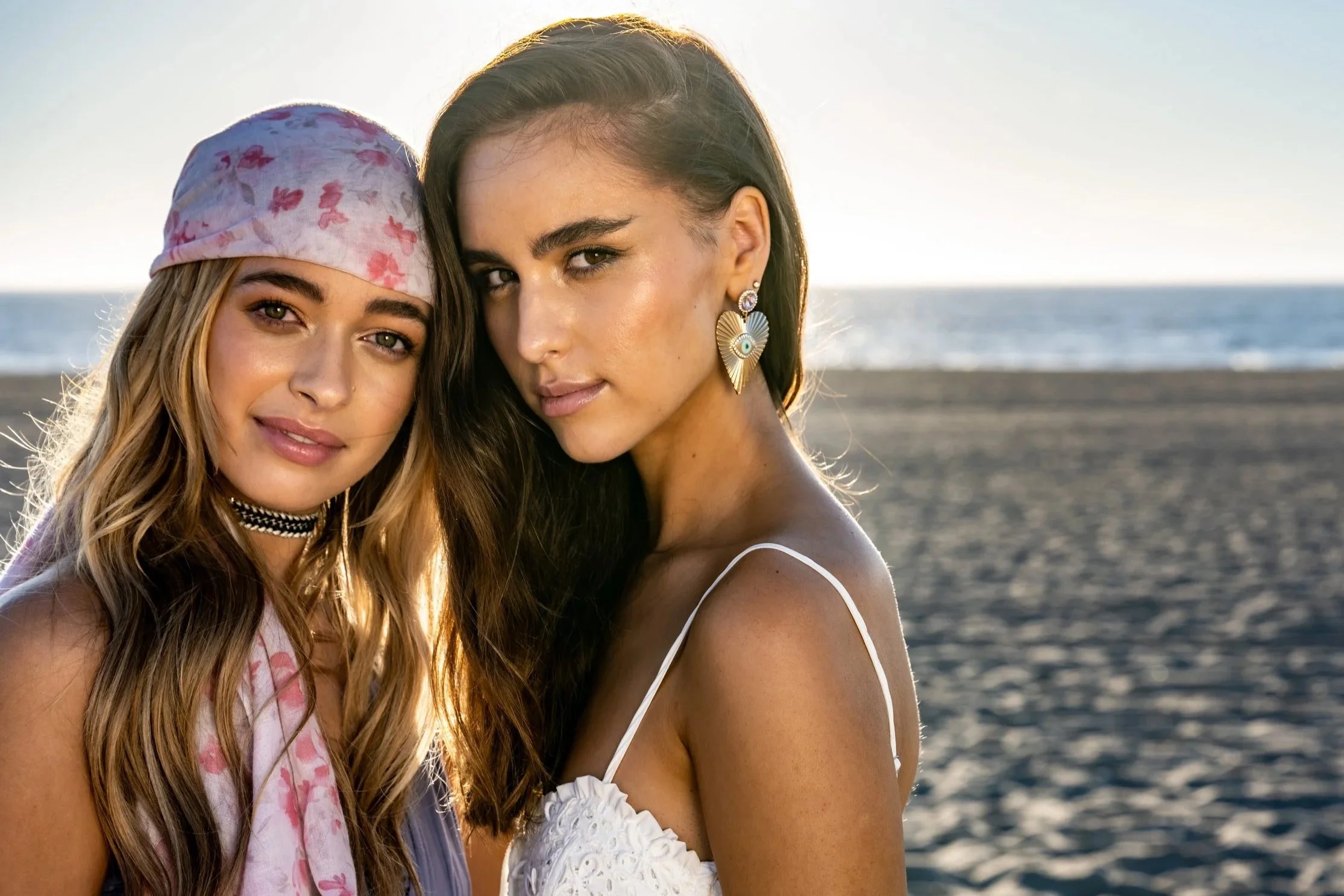 Two women standing close together on a beach at sunset, with the ocean in the background. They are both looking at the camera.
