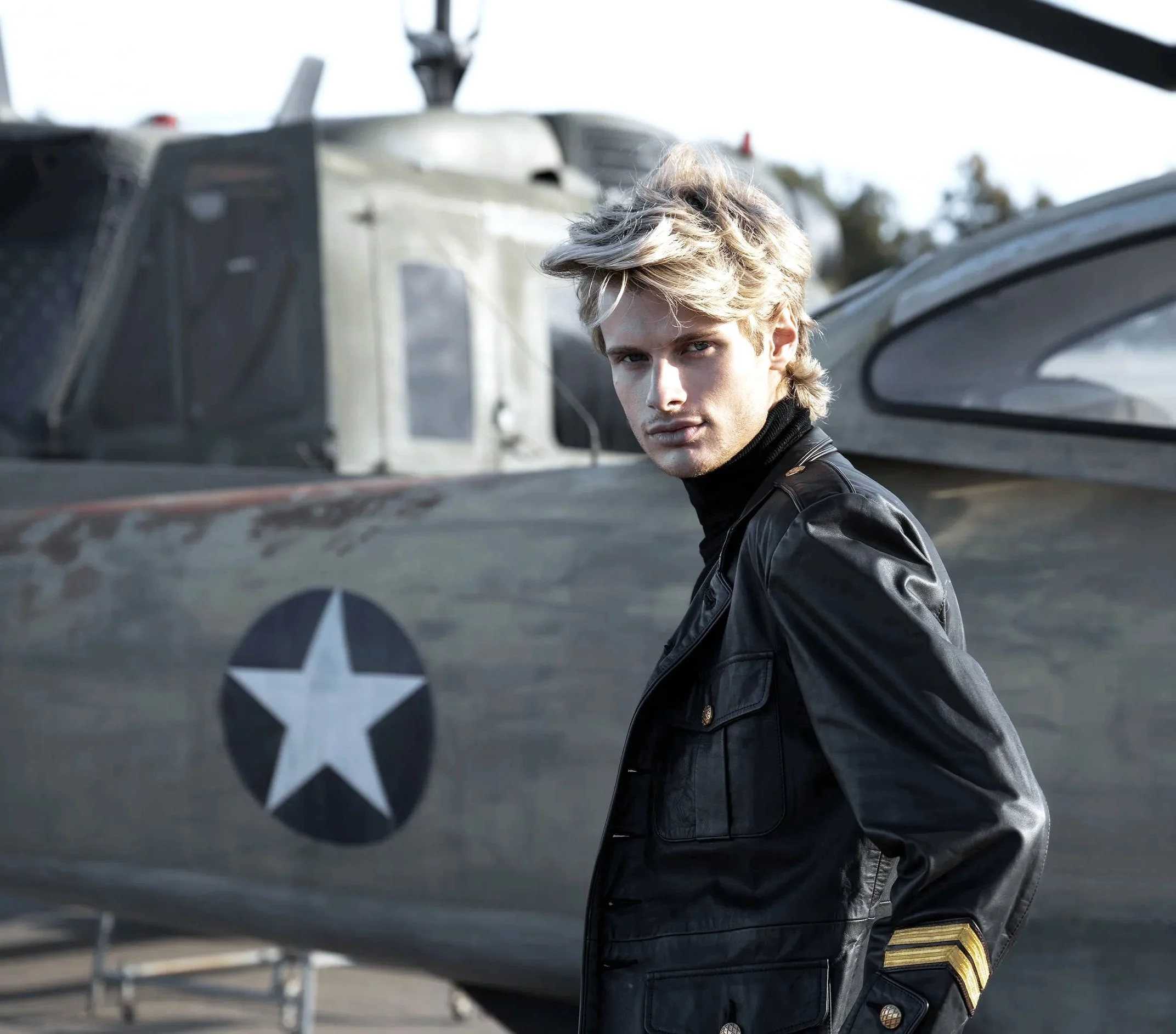 Young man with blonde hair and a leather jacket standing in front of a military helicopter with a U.S. star insignia.