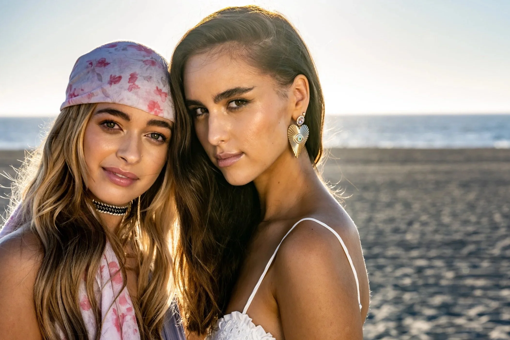 Two young women taking a selfie on the beach at sunset, one with a pink floral headscarf and choker, the other with earrings and a white spaghetti strap top.