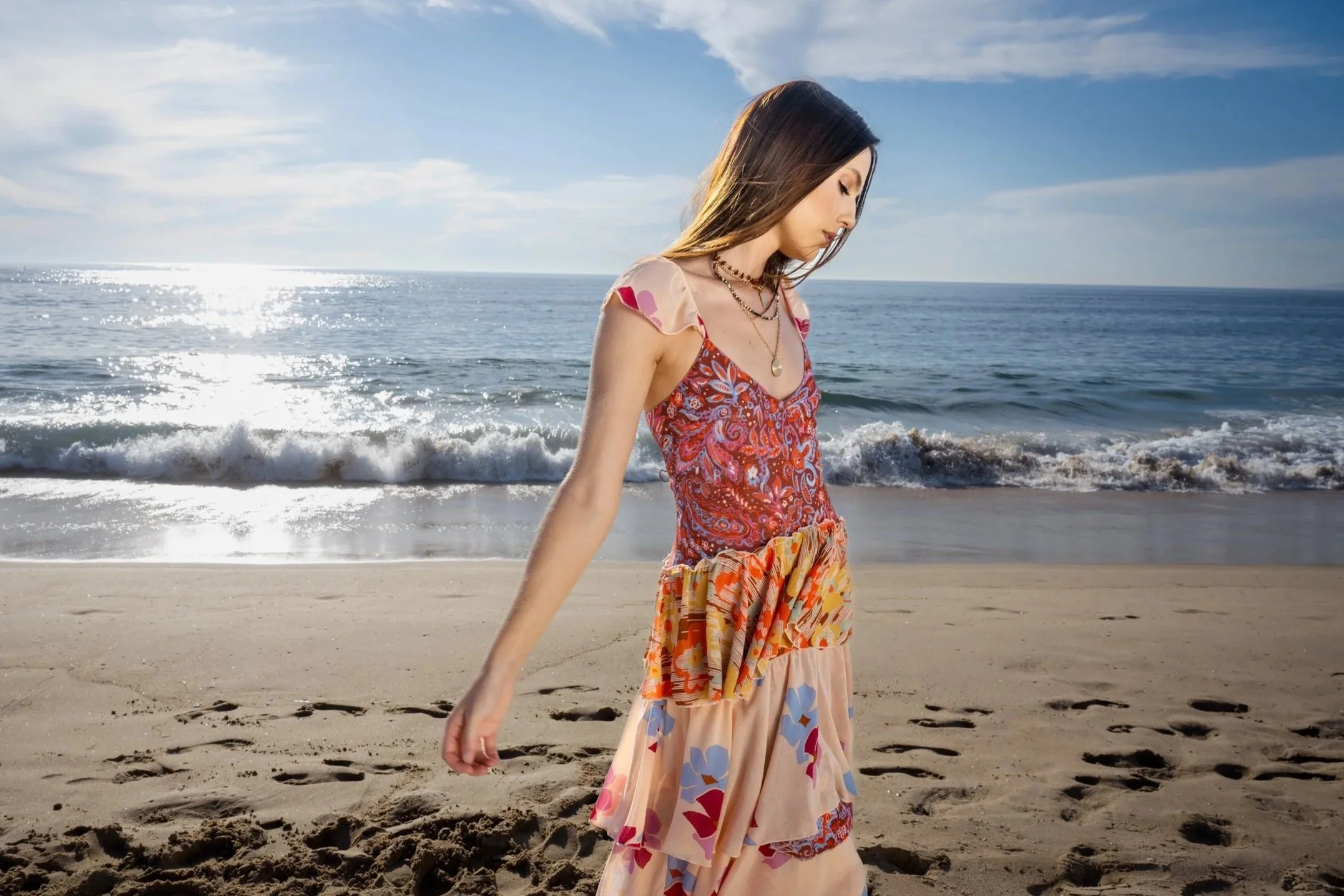 Woman walking on a sandy beach near the ocean with waves and a blue sky behind her.