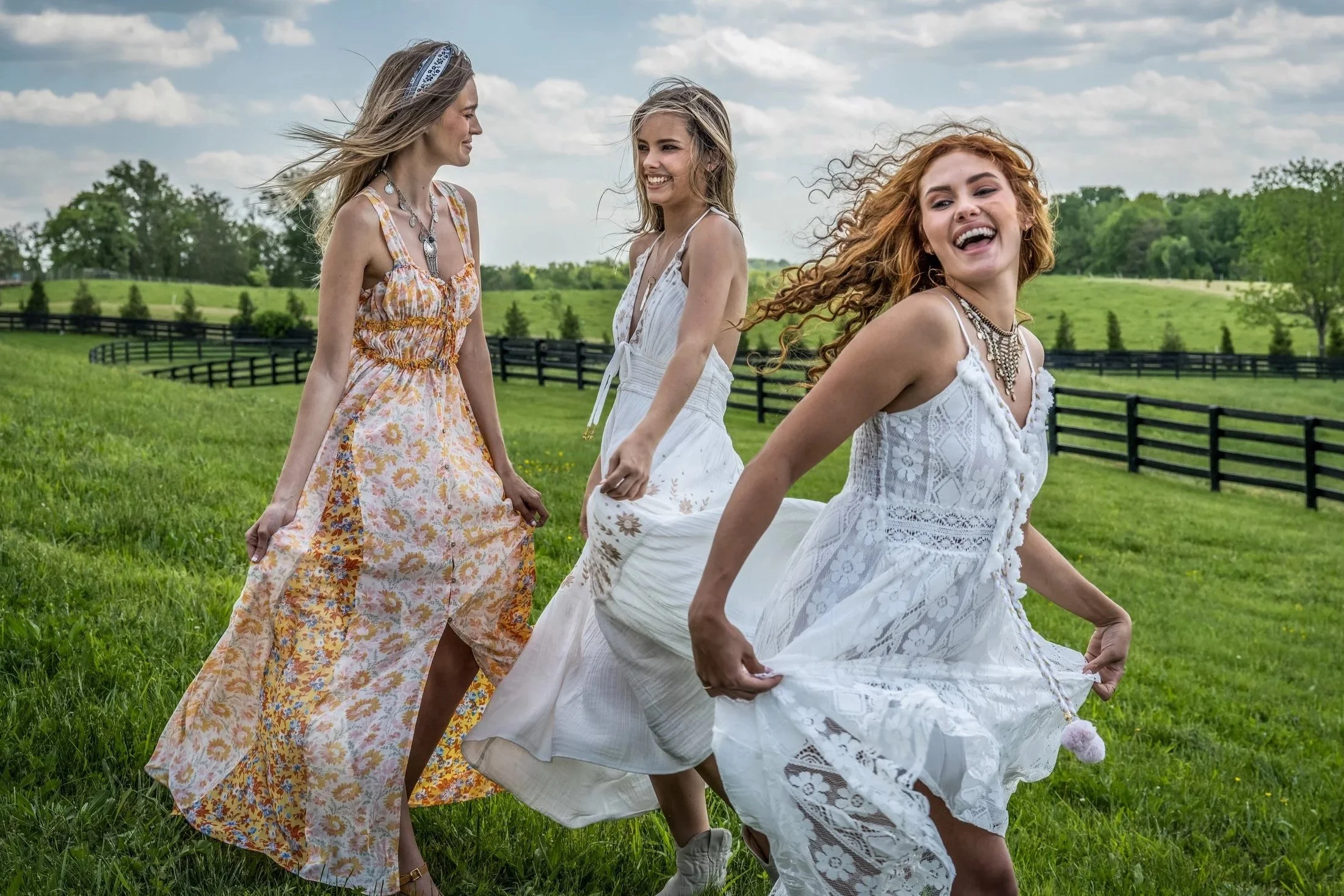 Four women in white and floral dresses walking and dancing in a green field with a black fence and trees in the background.
