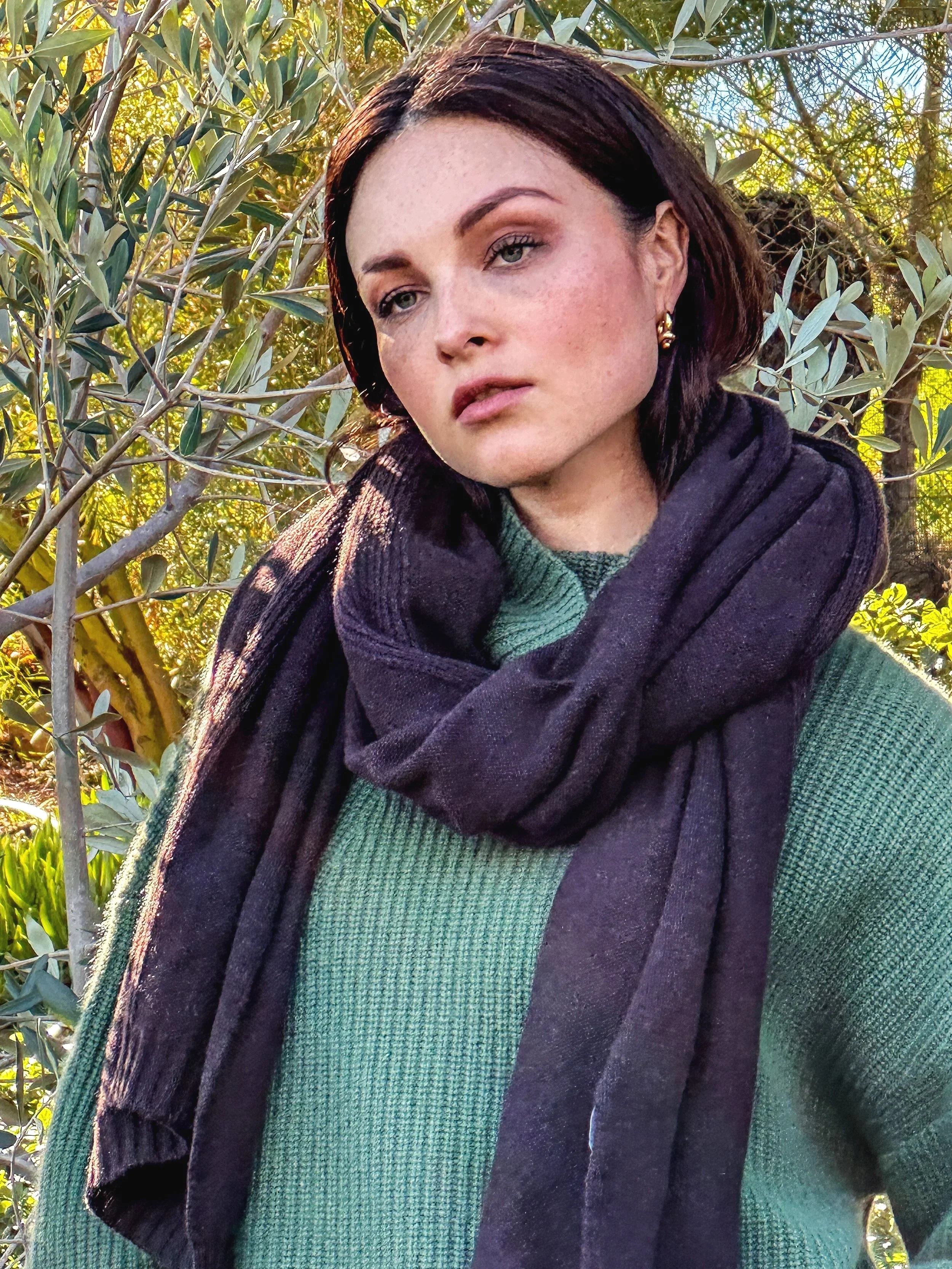 Woman with short dark hair standing outdoors among autumn trees, wearing a green sweater and black scarf.
