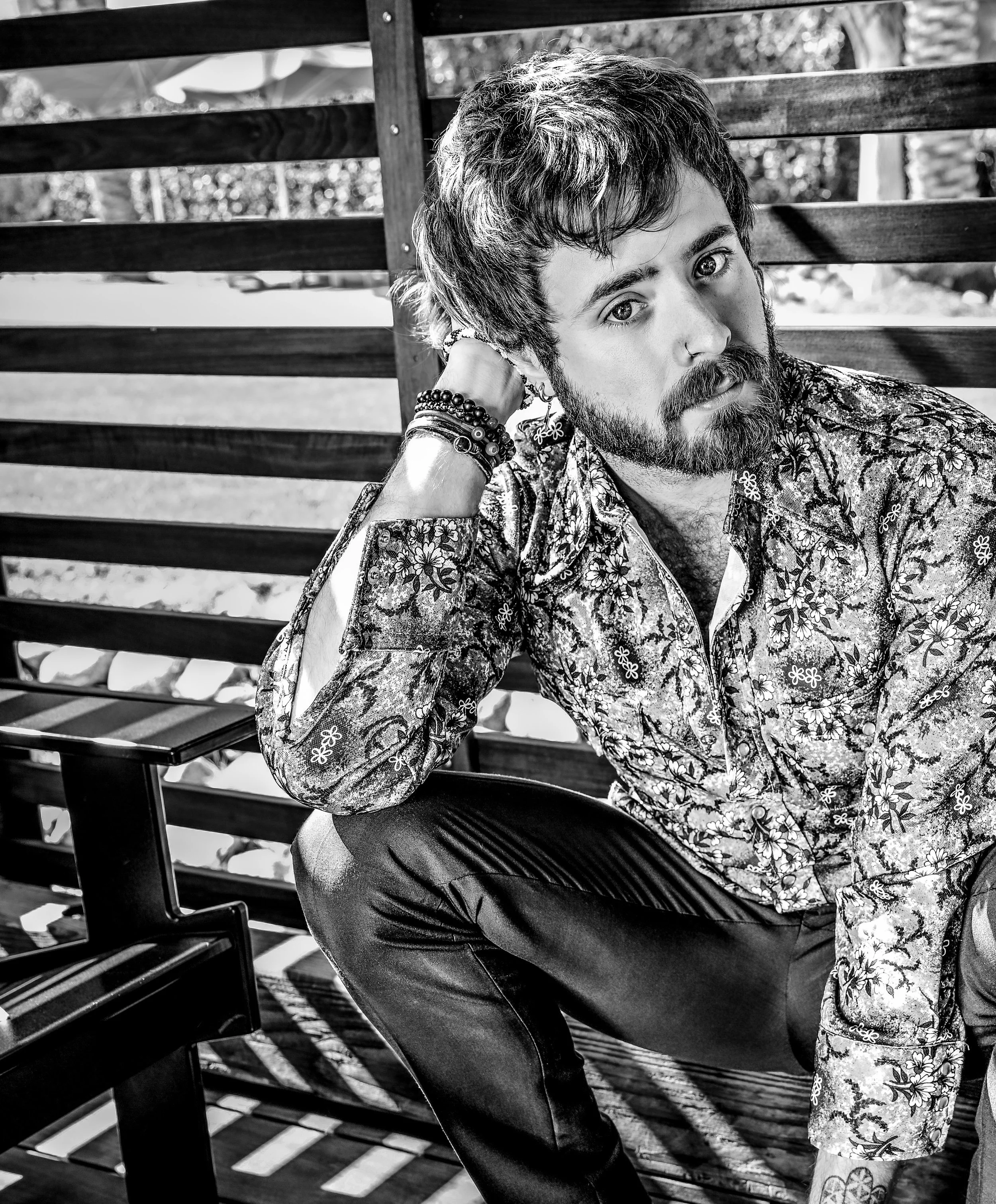 Black-and-white portrait of a man with curly hair and a beard sitting on a wooden bench and looking at the camera