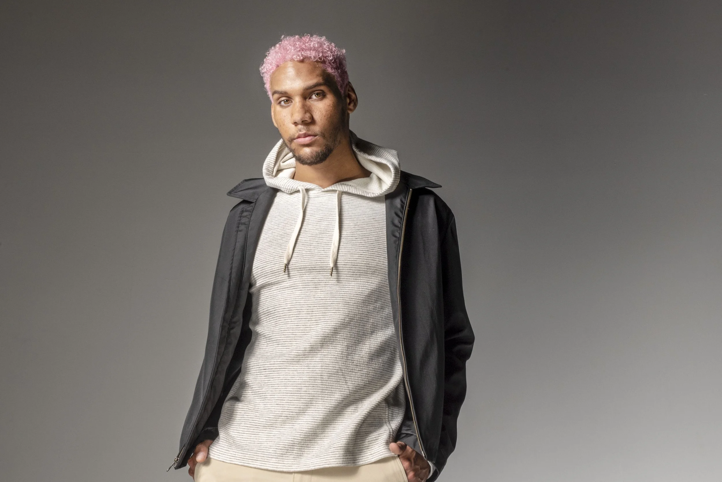 Man with pink curly hair wearing a black jacket over a light gray hoodie, standing against a plain gray background.
