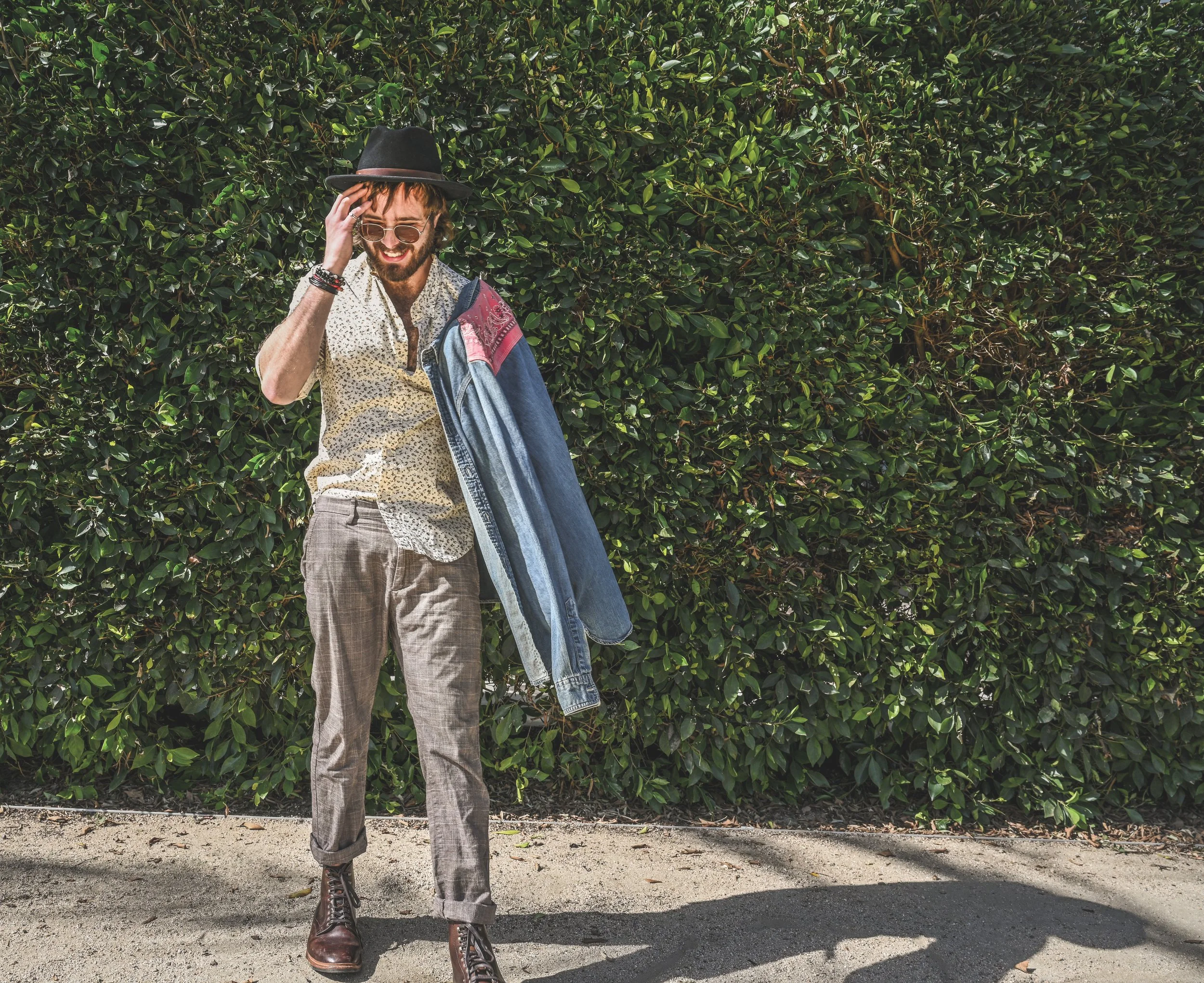 Man wearing a wide-brim hat and sunglasses standing in front of dense green bushes with a denim jacket over his shoulder.