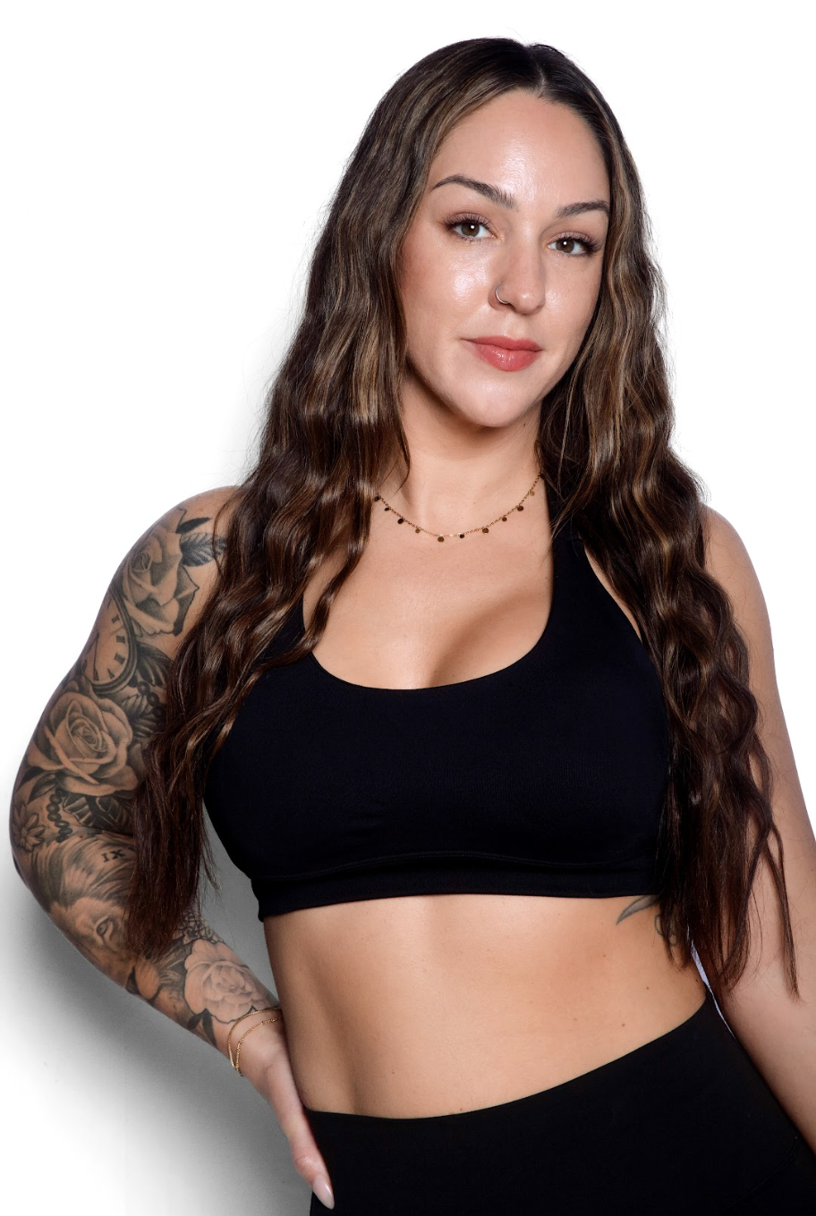 A woman with long wavy brown hair, a nose ring, and tattoos on her left arm, wearing a black sports bra and gold jewelry, posing against a white background.