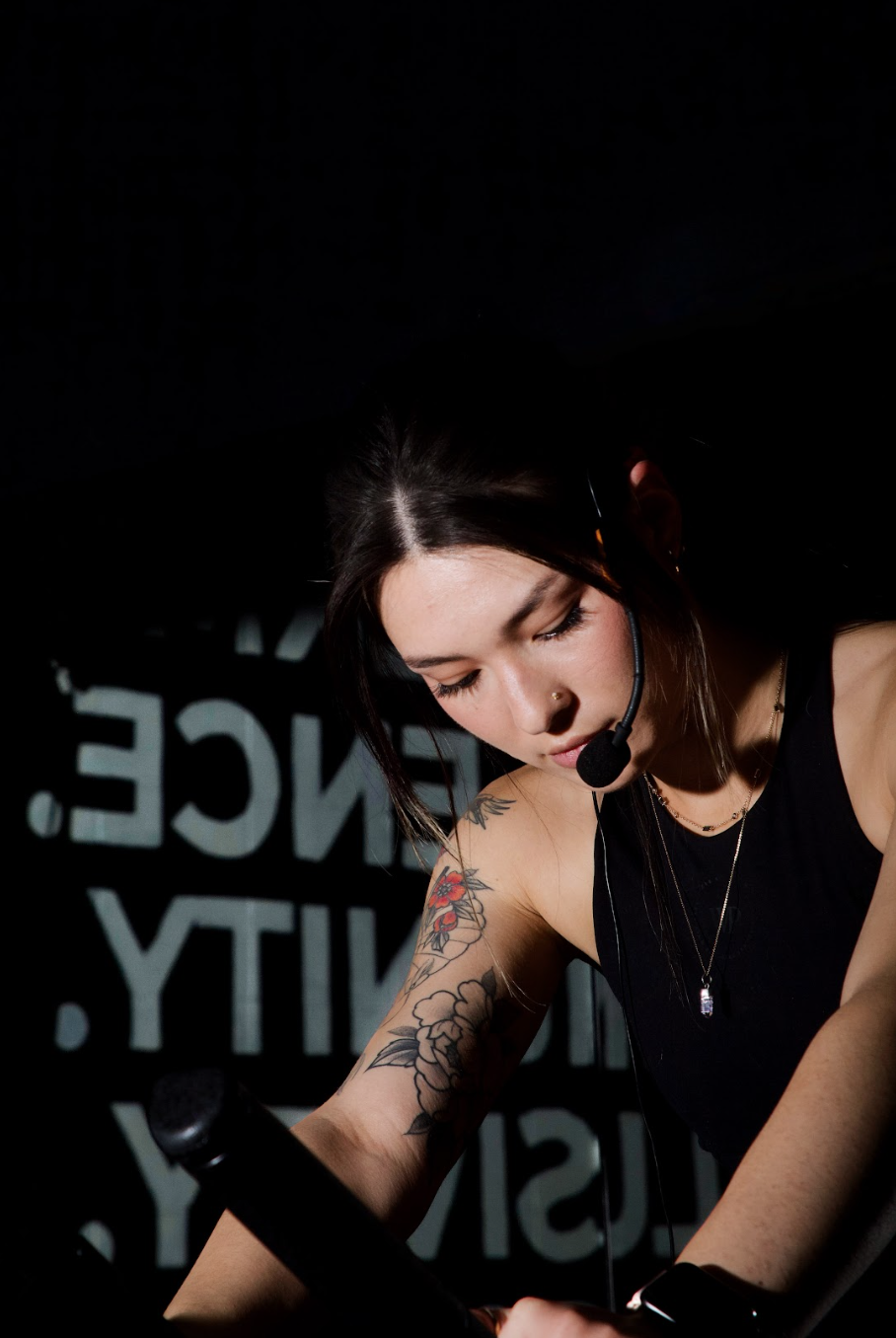 A woman with dark hair, tattoos on her arm, and a nose piercing, wearing a headset with microphone, looking downward while working or speaking.