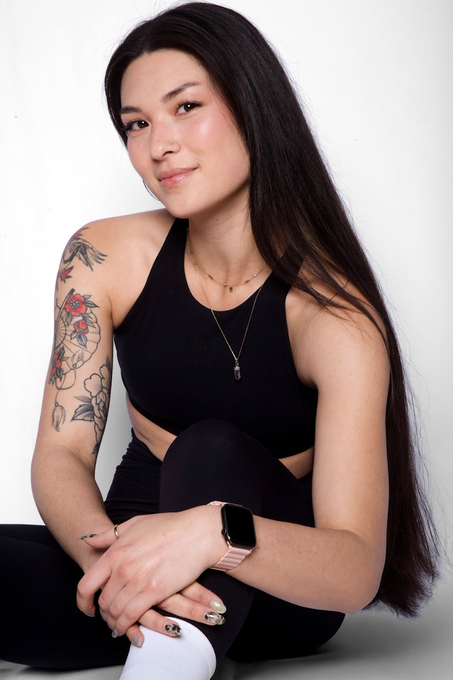 Young woman with long dark hair, tattoos on her left arm, wearing a black sleeveless top, black leggings, white socks, a smartwatch, and layered necklaces, sitting against a white background.